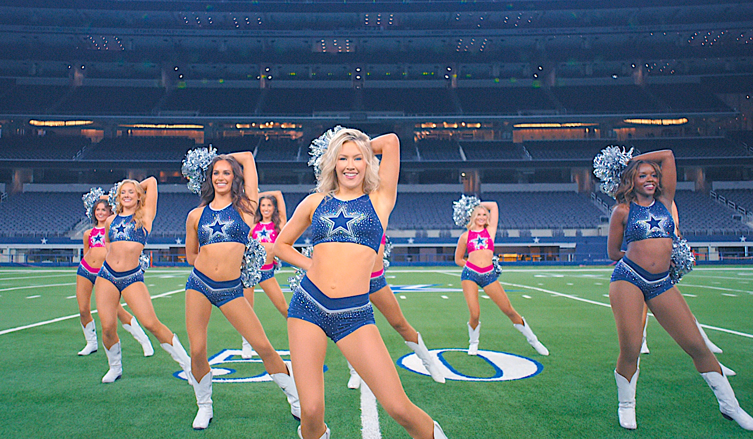 Cheerleaders perform a choreographed dance on a football field, wearing coordinated, star-themed outfits with pom-poms