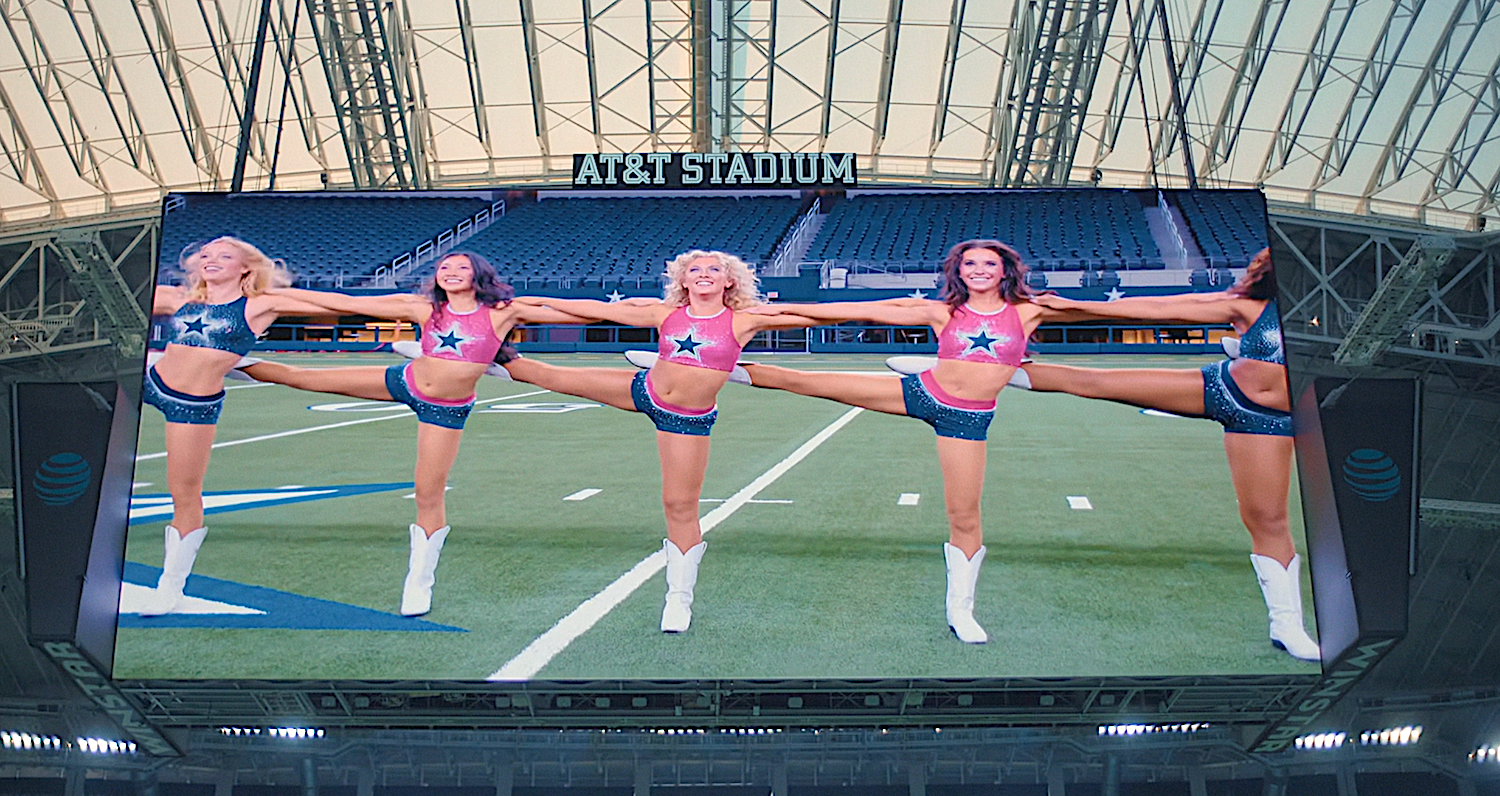Cheerleaders perform high kicks on a giant screen at AT&T Stadium. They wear coordinated outfits with star designs and boots