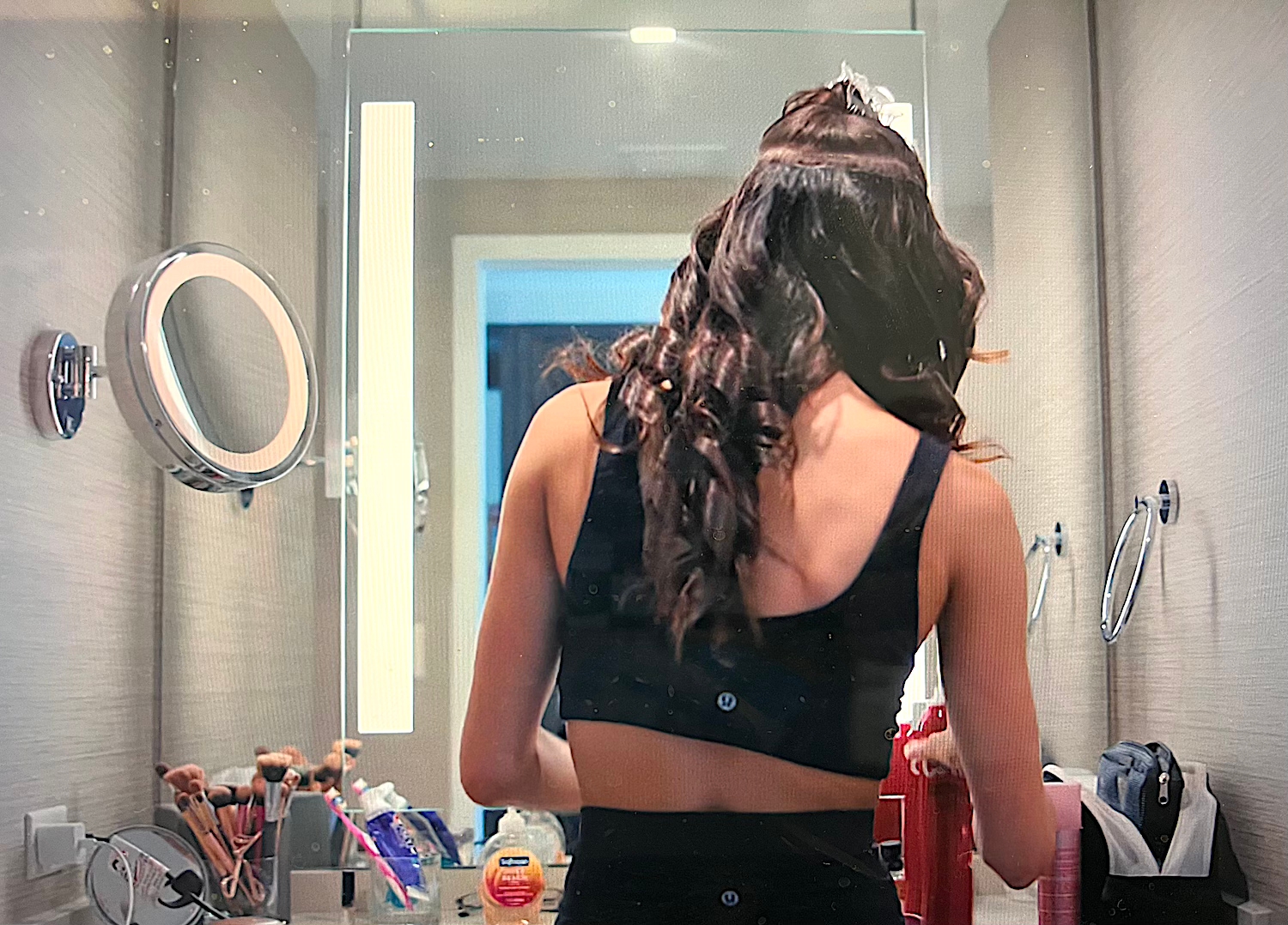 Person with wavy hair styling in a bathroom, wearing a sleeveless top. Bathroom counter has beauty products and a lighted mirror
