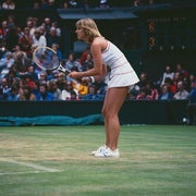 Tennis player in a focused stance on grass court during a match, wearing a classic white tennis outfit and holding a racquet. Crowd watches