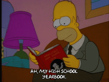 Homer Simpson, in a suit, sits on a couch and looks at a yearbook, saying, &quot;Ah, my high school yearbook.&quot;