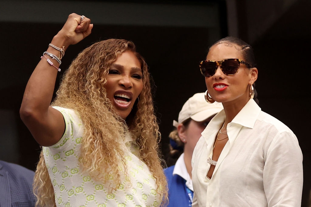 Two women at an event; one raises her fist and smiles. They wear stylish outfits and sunglasses