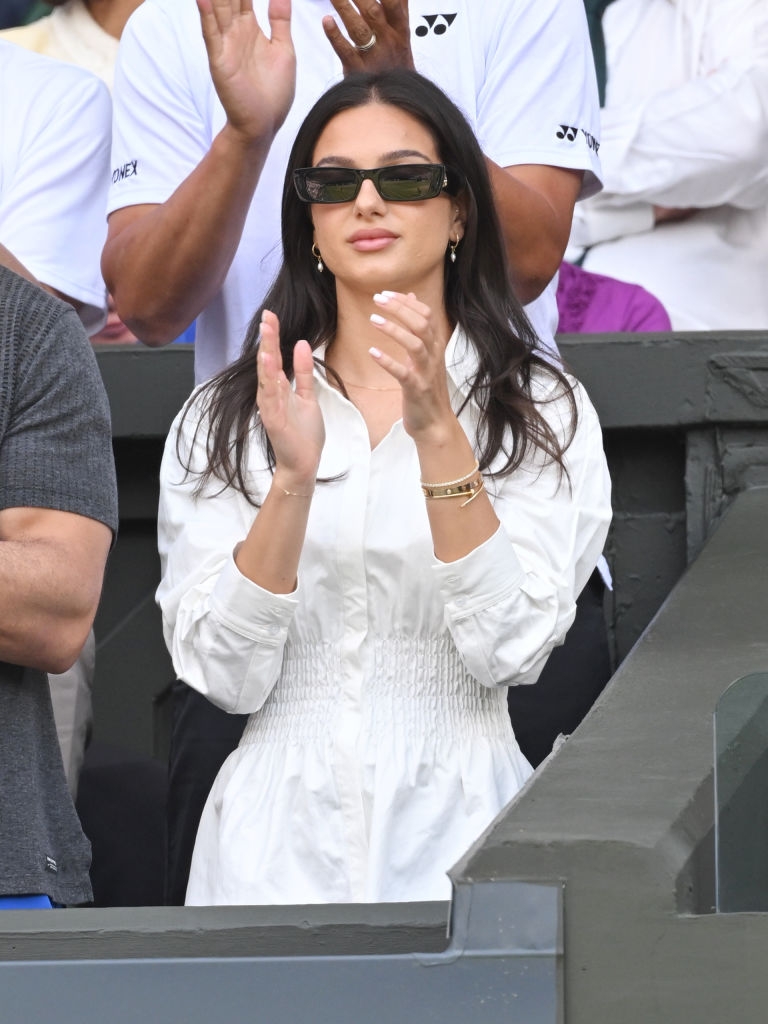 Person in stylish white dress and sunglasses applauding at an event; background shows casually dressed crowd