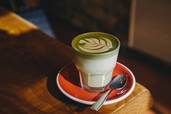 A glass of matcha latte with leaf latte art on a red saucer, placed on a wooden table with a spoon beside it