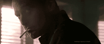 Person with cigarette looks over shoulder in dim light; text warns about smoking&#x27;s health risks