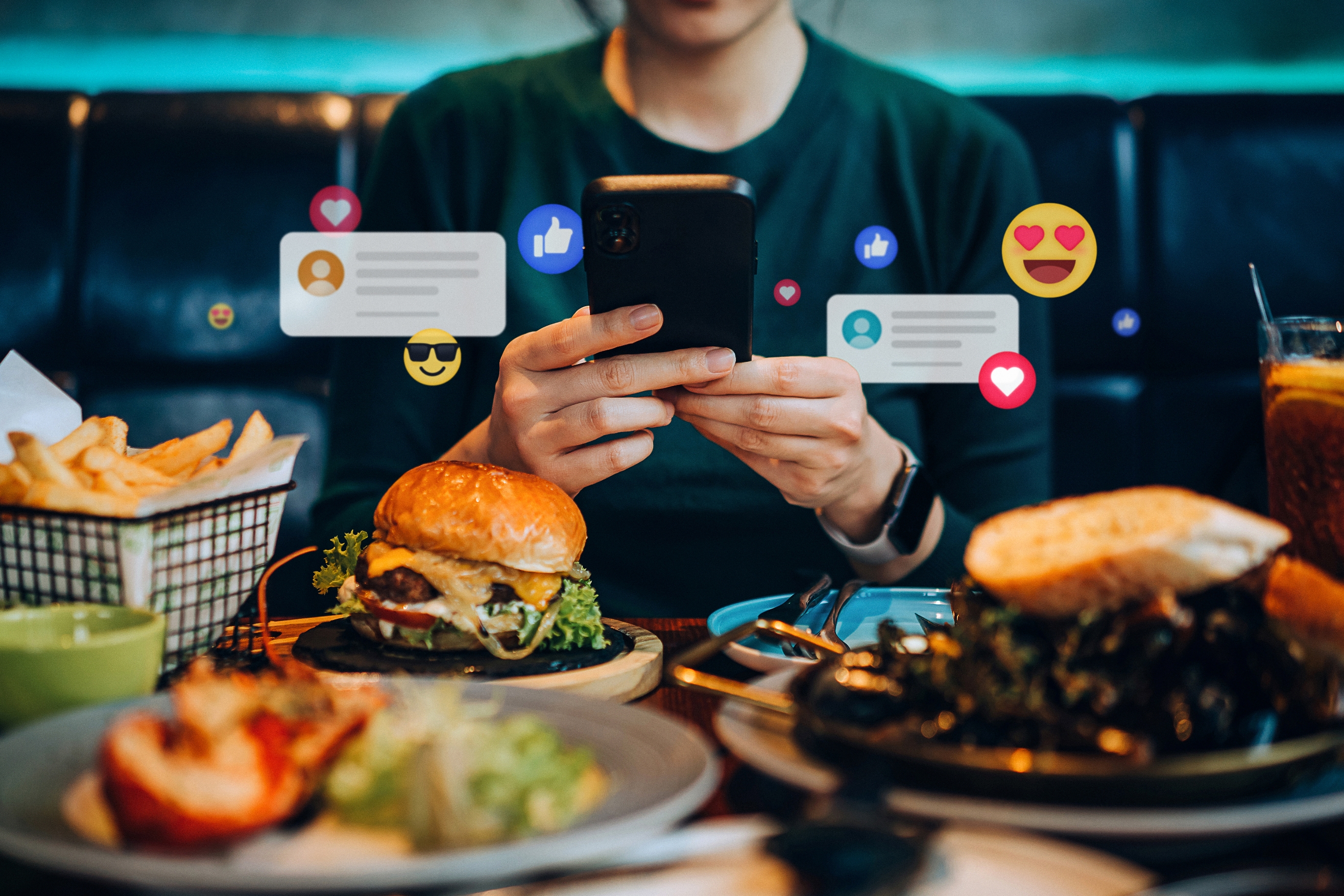 Person at table using smartphone surrounded by plates of food; digital social media icons hover above