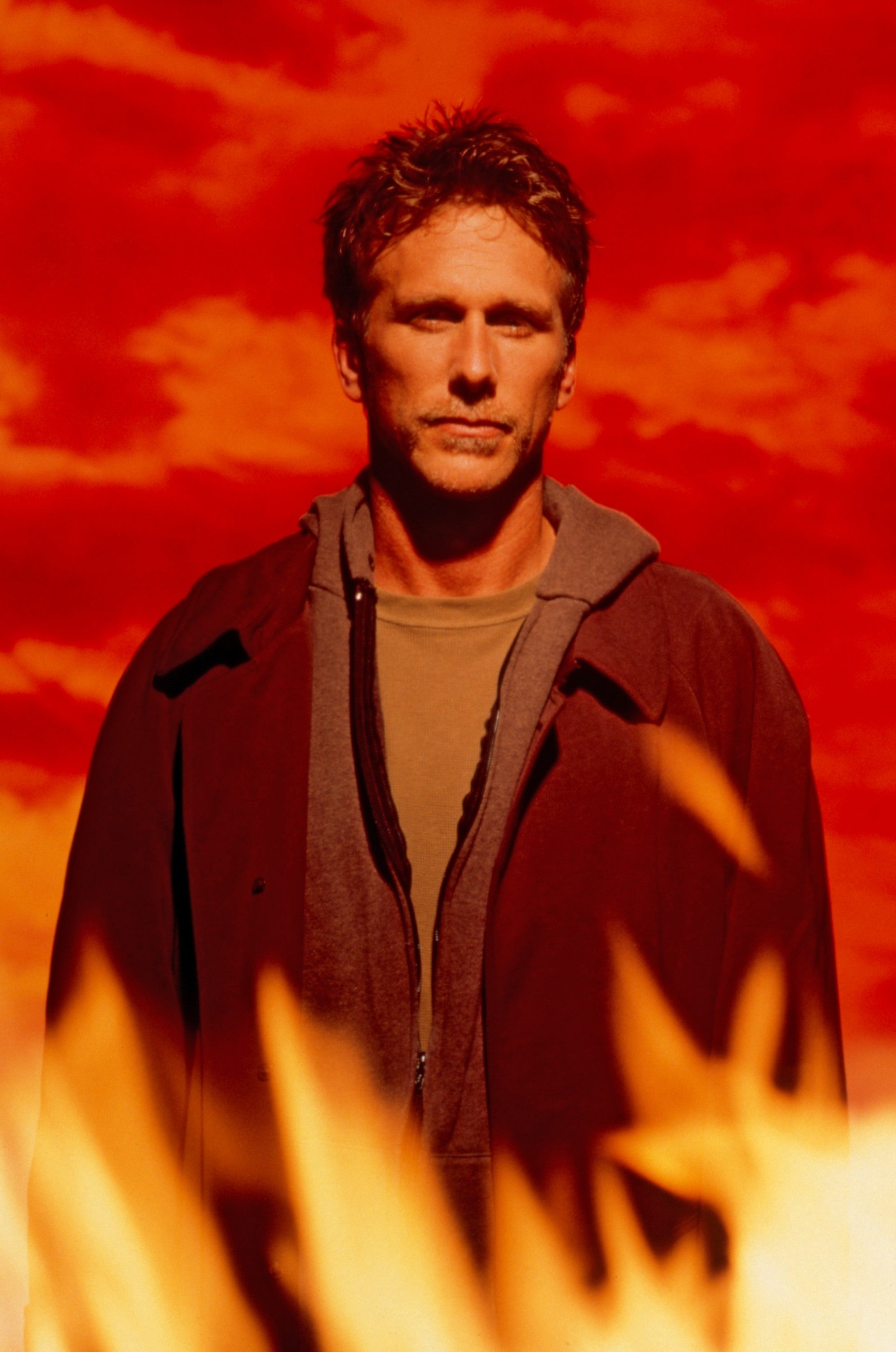 Person stands against a fiery red background, wearing a jacket over a shirt, looking determined