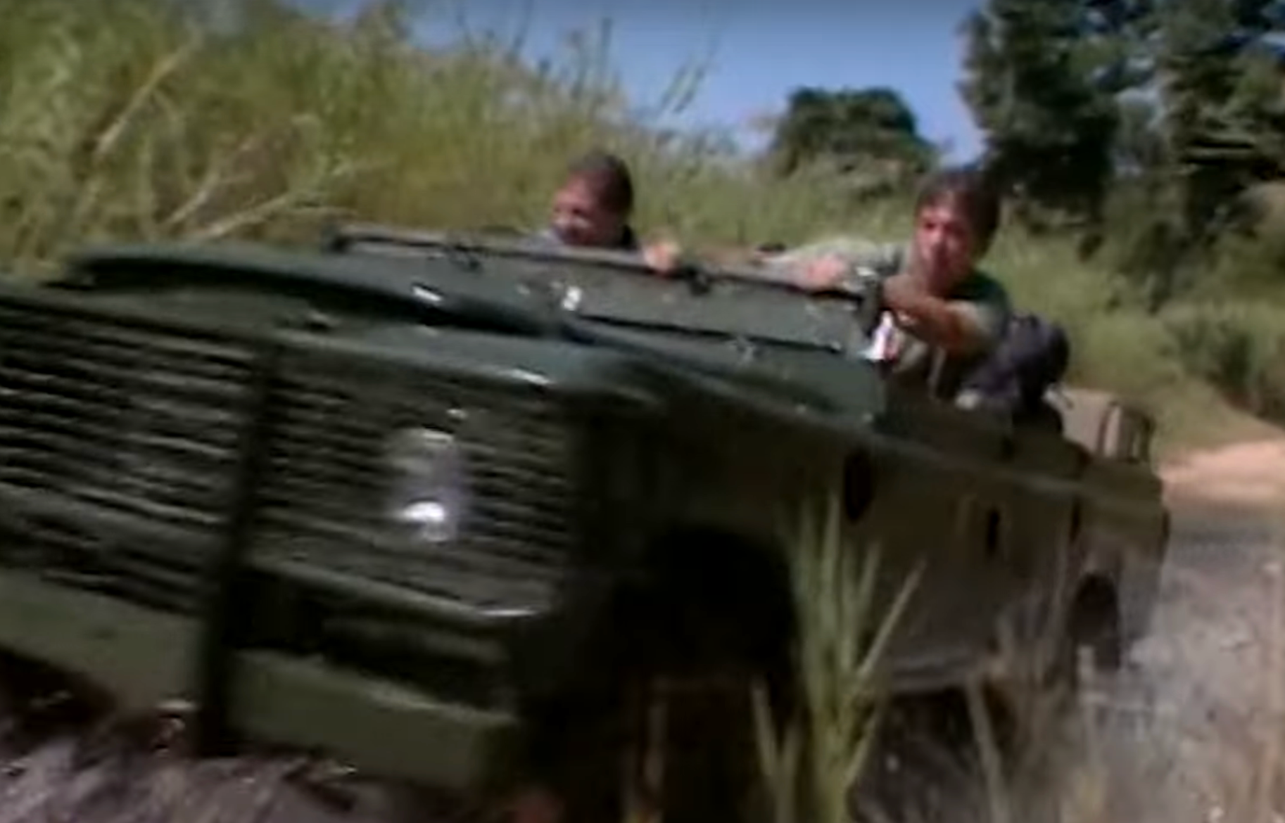 Two people drive a rugged off-road vehicle through lush terrain, appearing focused and adventurous