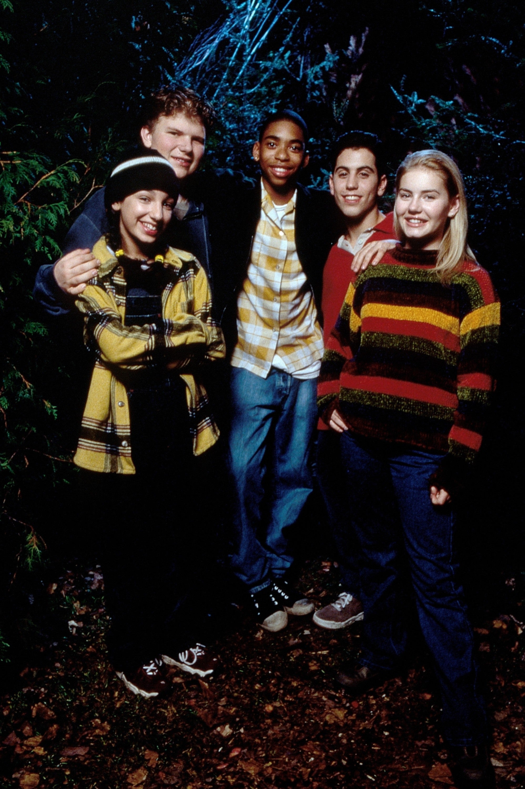 Group of five young adults in casual, layered clothing stand closely together, smiling, outdoors at night