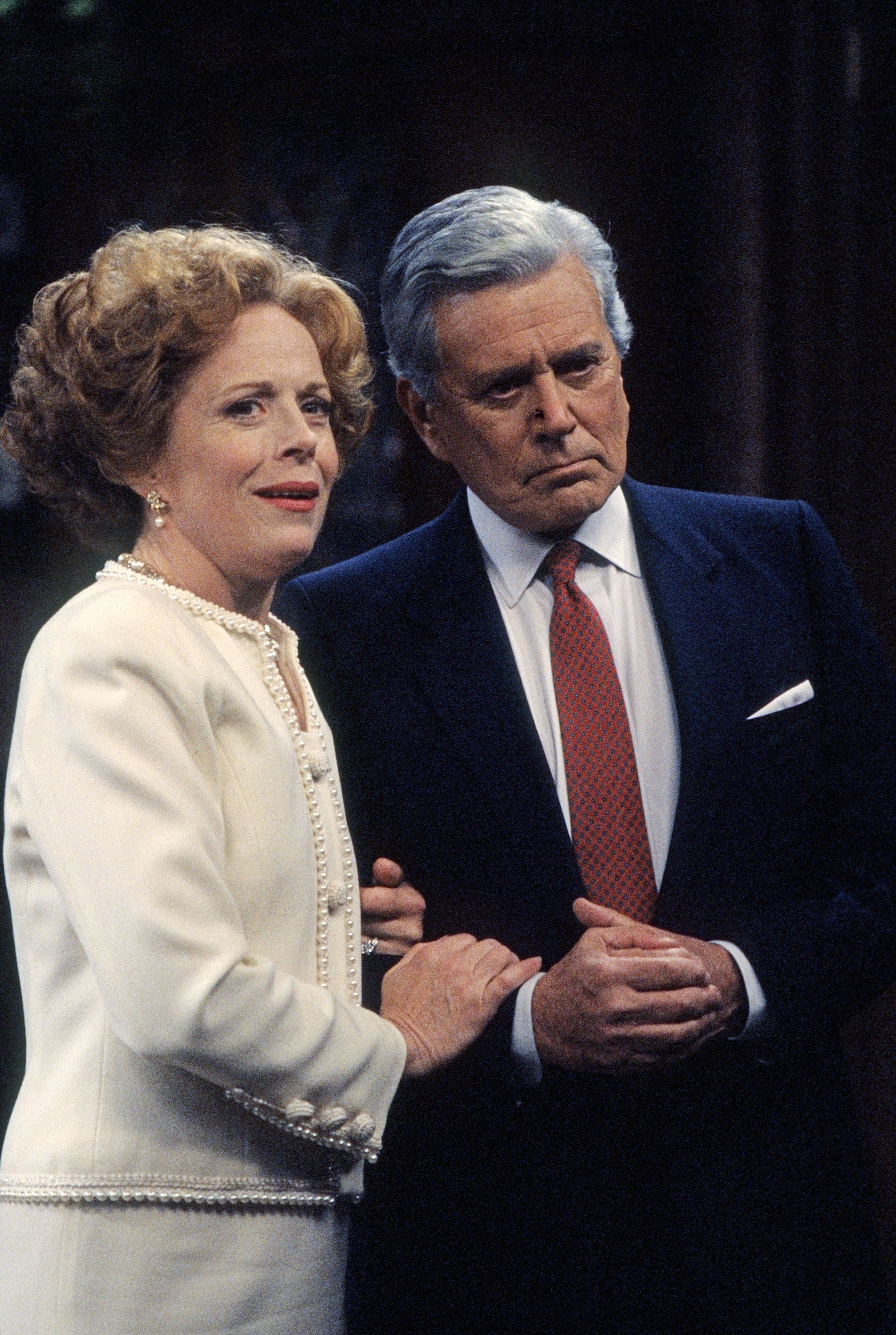 Two actors in formal 1980s attire stand closely, portraying an intense scene, suggesting drama in a TV or movie setting