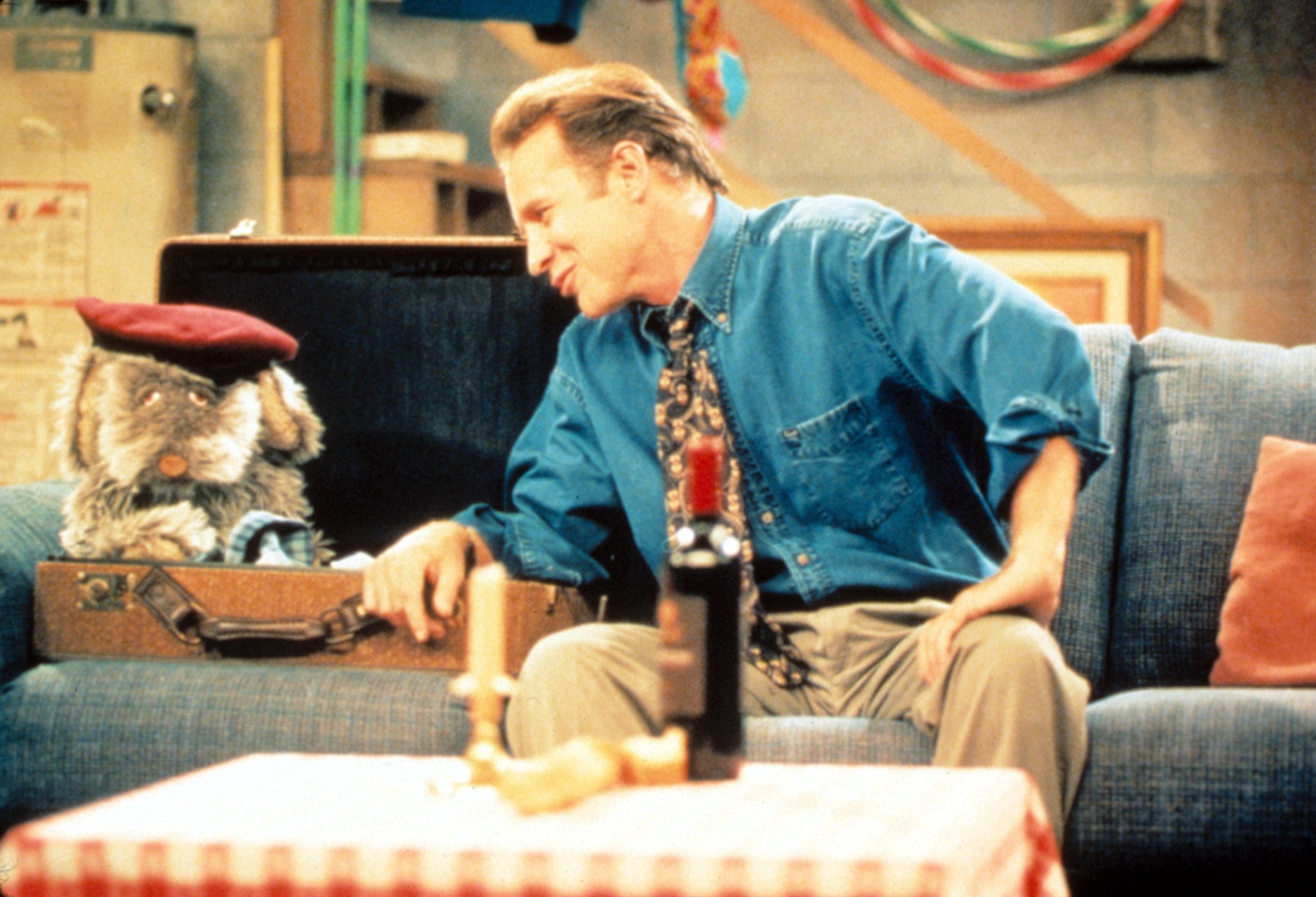 A man in a blue shirt and tie talks animatedly with a puppet sitting inside an open suitcase on a living room set