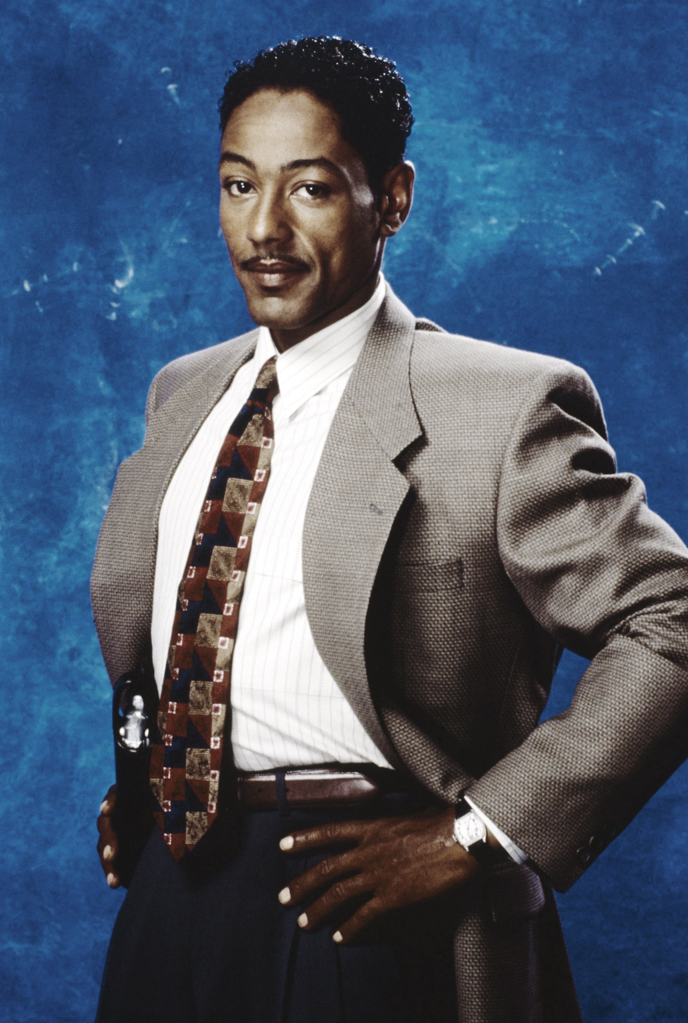 Person in a suit and tie posing against a plain backdrop, hands on hips