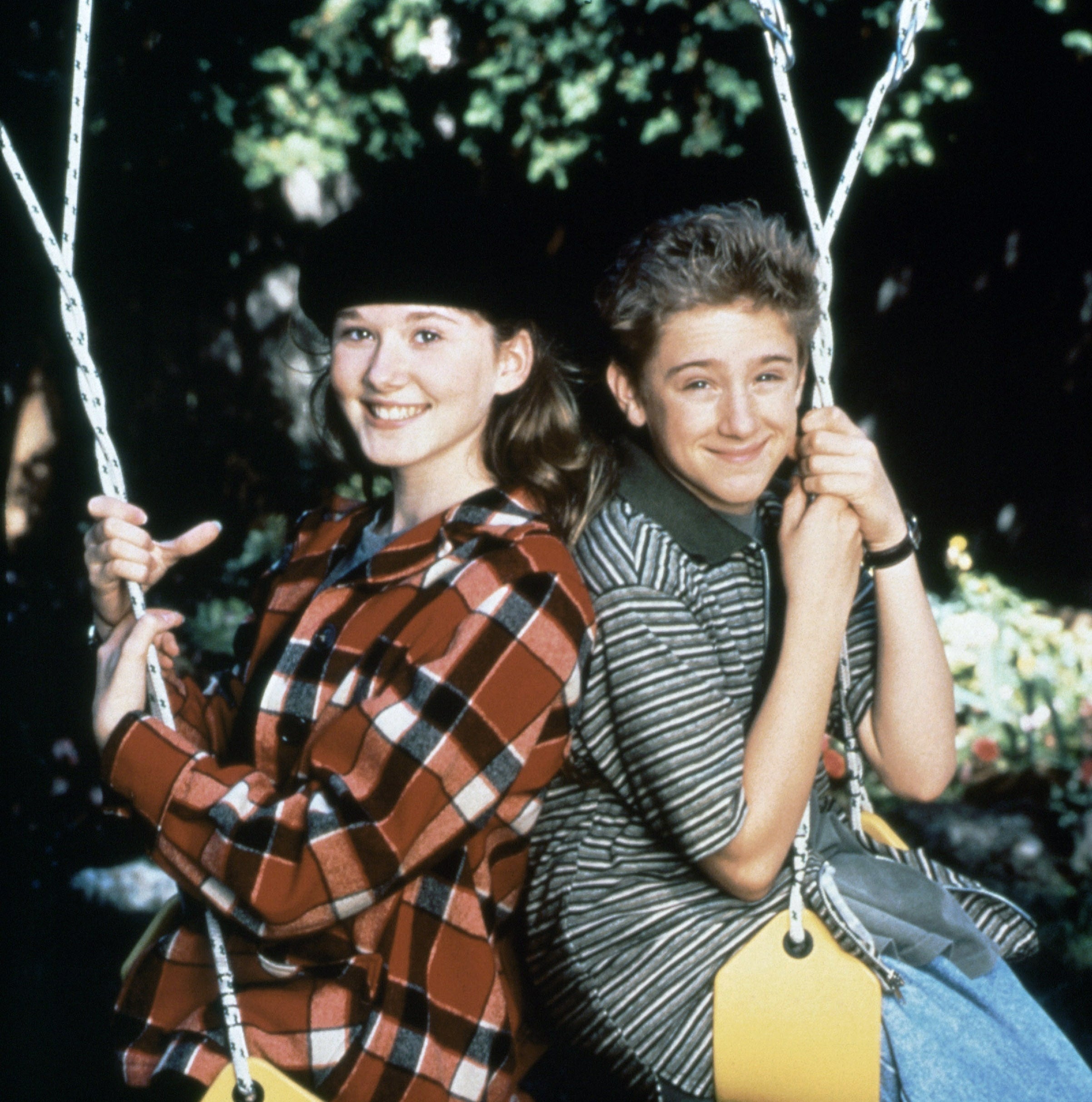 Two young actors are sitting on swings, smiling. One is wearing a plaid jacket and hat, the other a striped shirt. The setting is outdoors