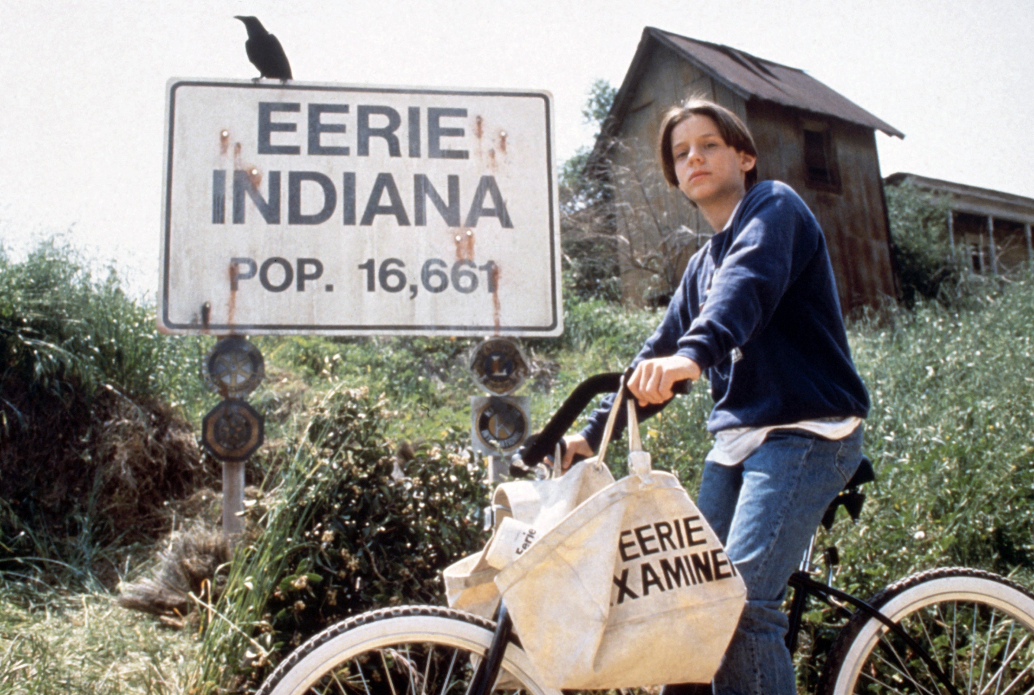 Person on a bike with a newspaper bag stands by Eerie Indiana sign, pop. 16,661, with a crow perched on top