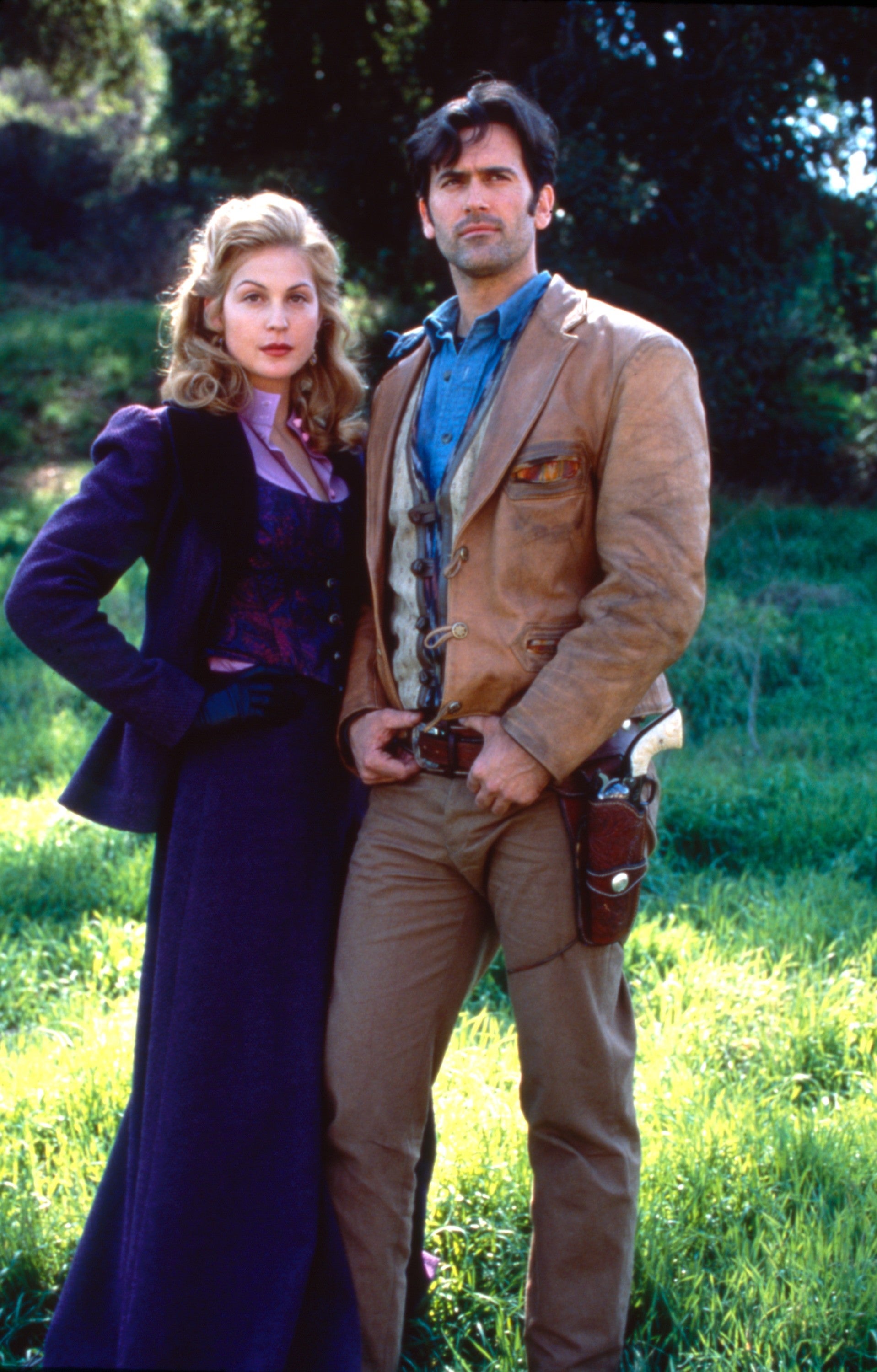 Two actors in vintage Western attire stand in a grassy outdoor setting. The woman wears a long coat, and the man is in a suede jacket with a holster