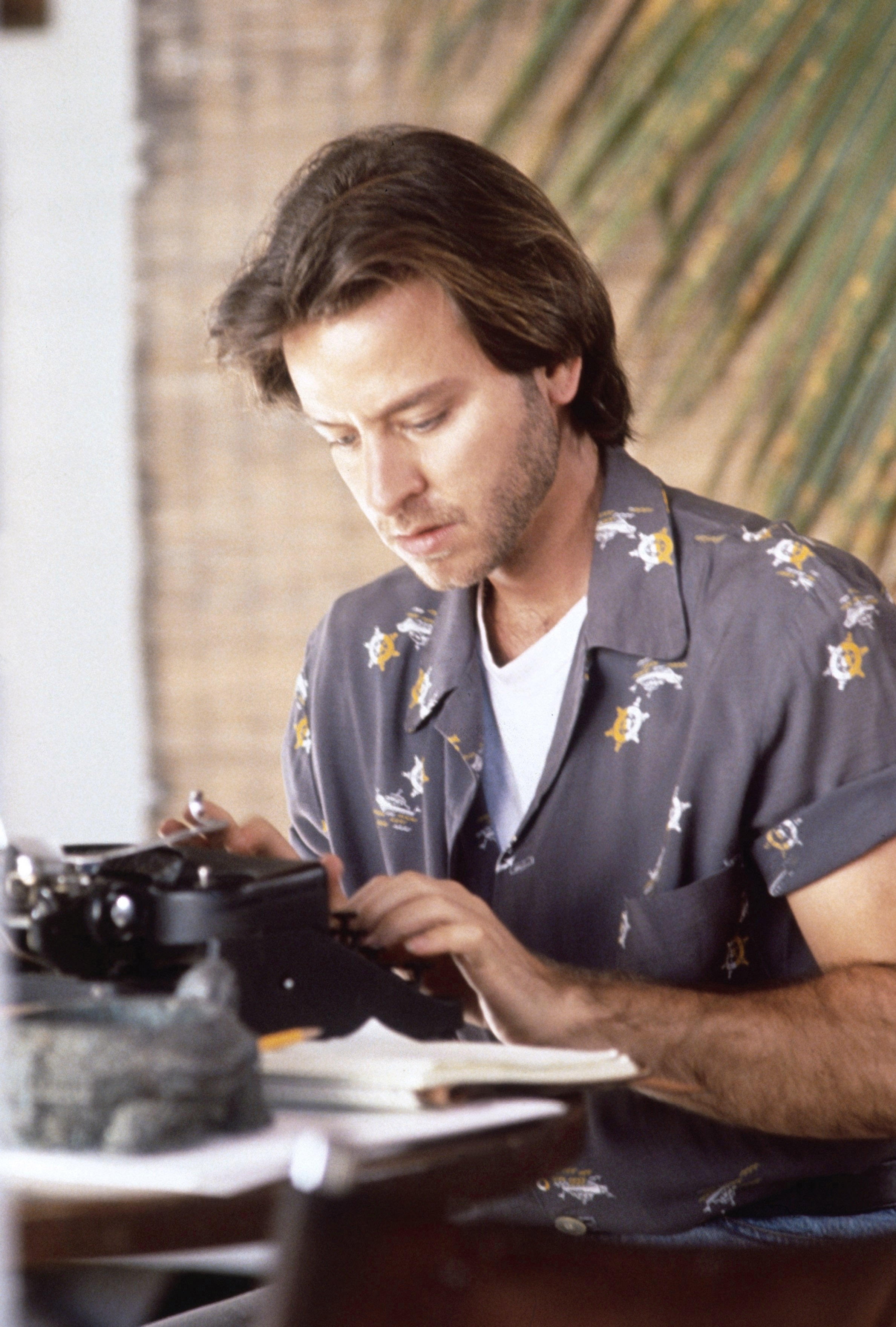 A person typing on a typewriter, wearing a patterned shirt with a thoughtful expression