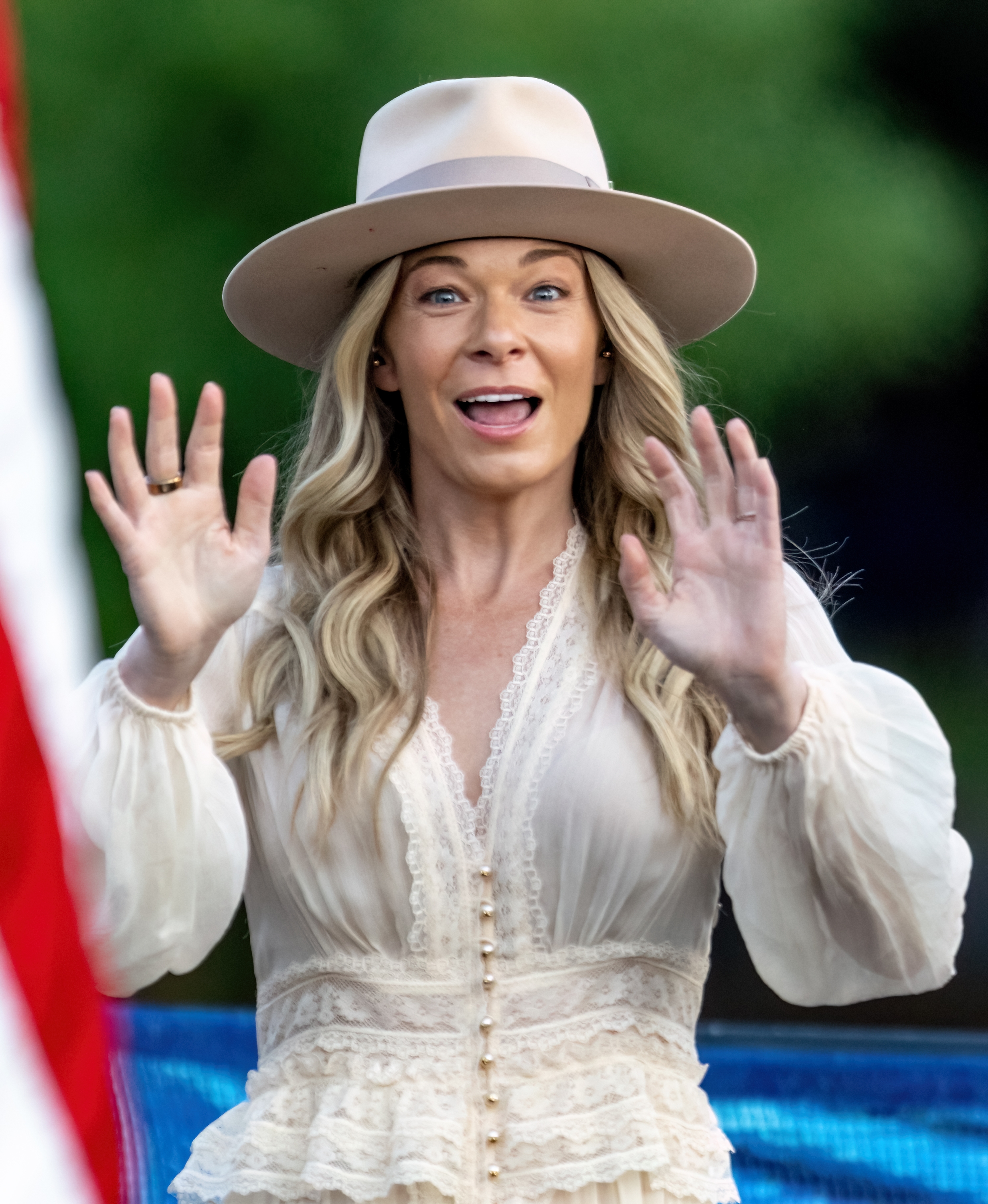 LeAnn Rimes wearing a wide-brim hat and a lace dress, with arms raised in excitement, standing outdoors