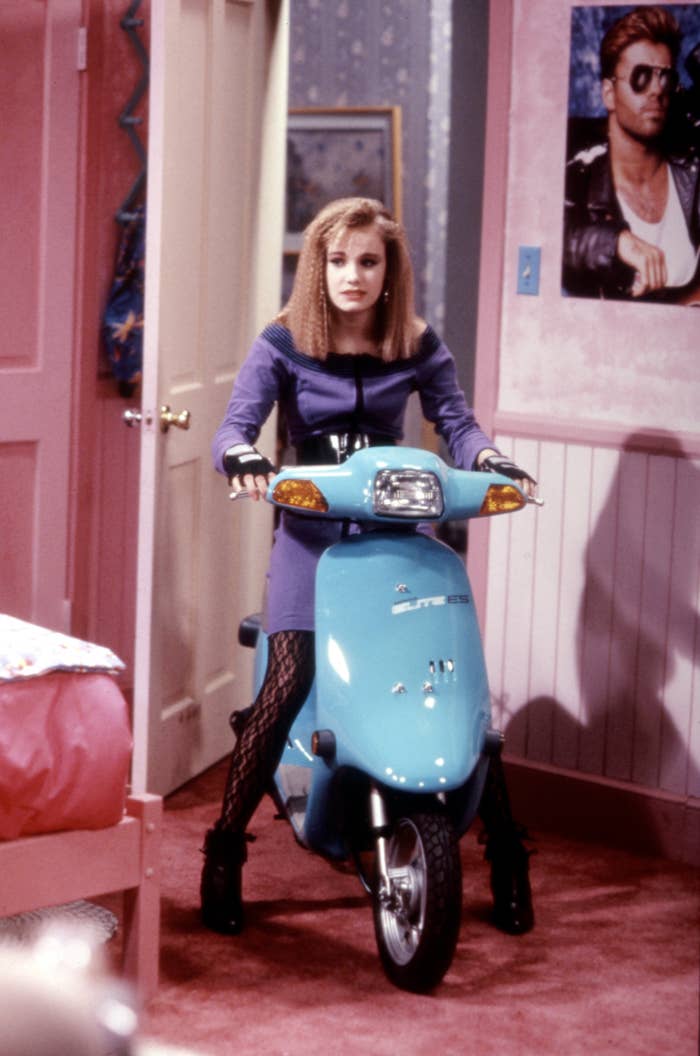 Person in '90s fashion stands in a room on a scooter, with a poster visible on the wall behind them