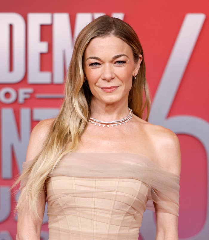 LeAnn Rimes on red carpet in off-the-shoulder gown with layered necklace, smiling in front of large event logo