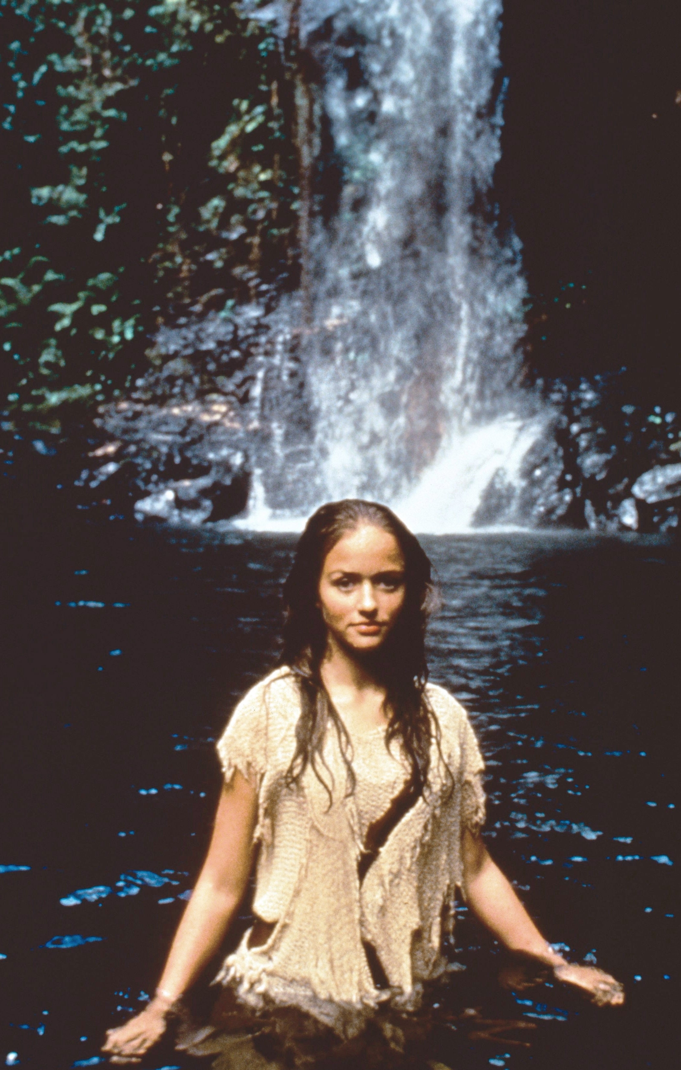Person standing in water near a waterfall, wearing a textured top, with a serene expression. Forest backdrop suggests a nature scene