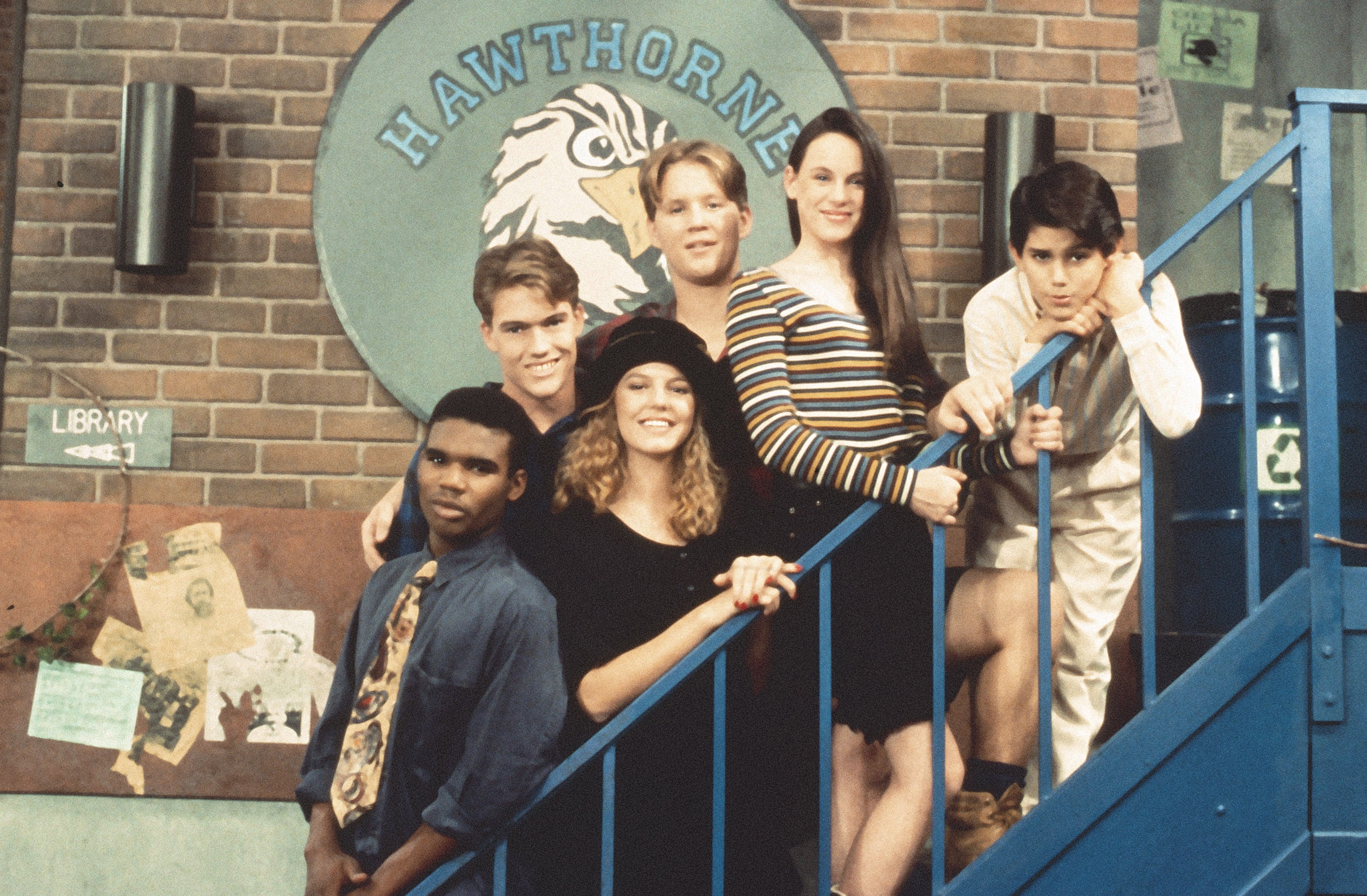 Group of young people from a 90s TV show posing on a staircase, smiling in casual, vintage clothing