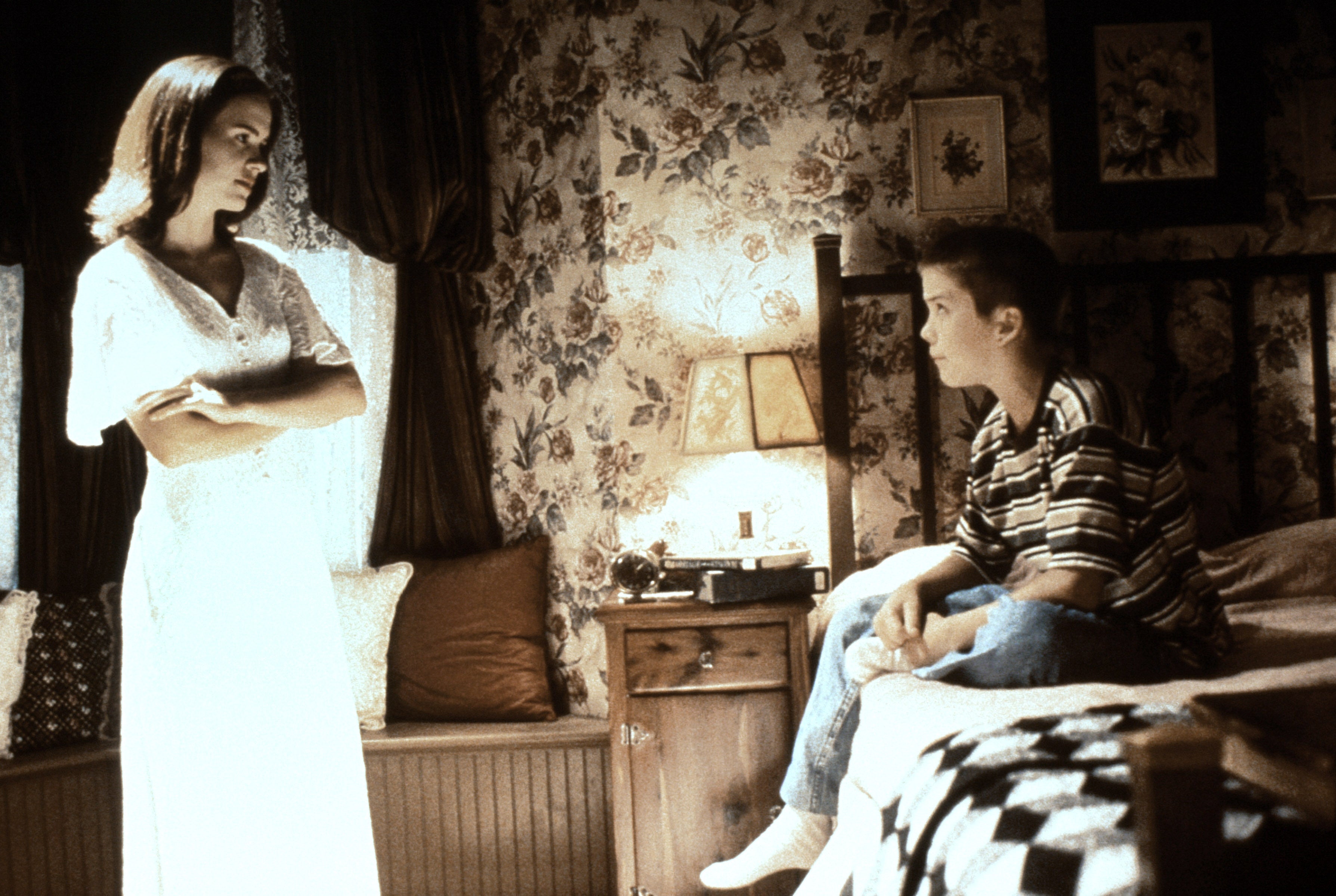 A woman in a white dress stands next to a bed, talking to a seated boy in a striped shirt in a floral wallpapered room