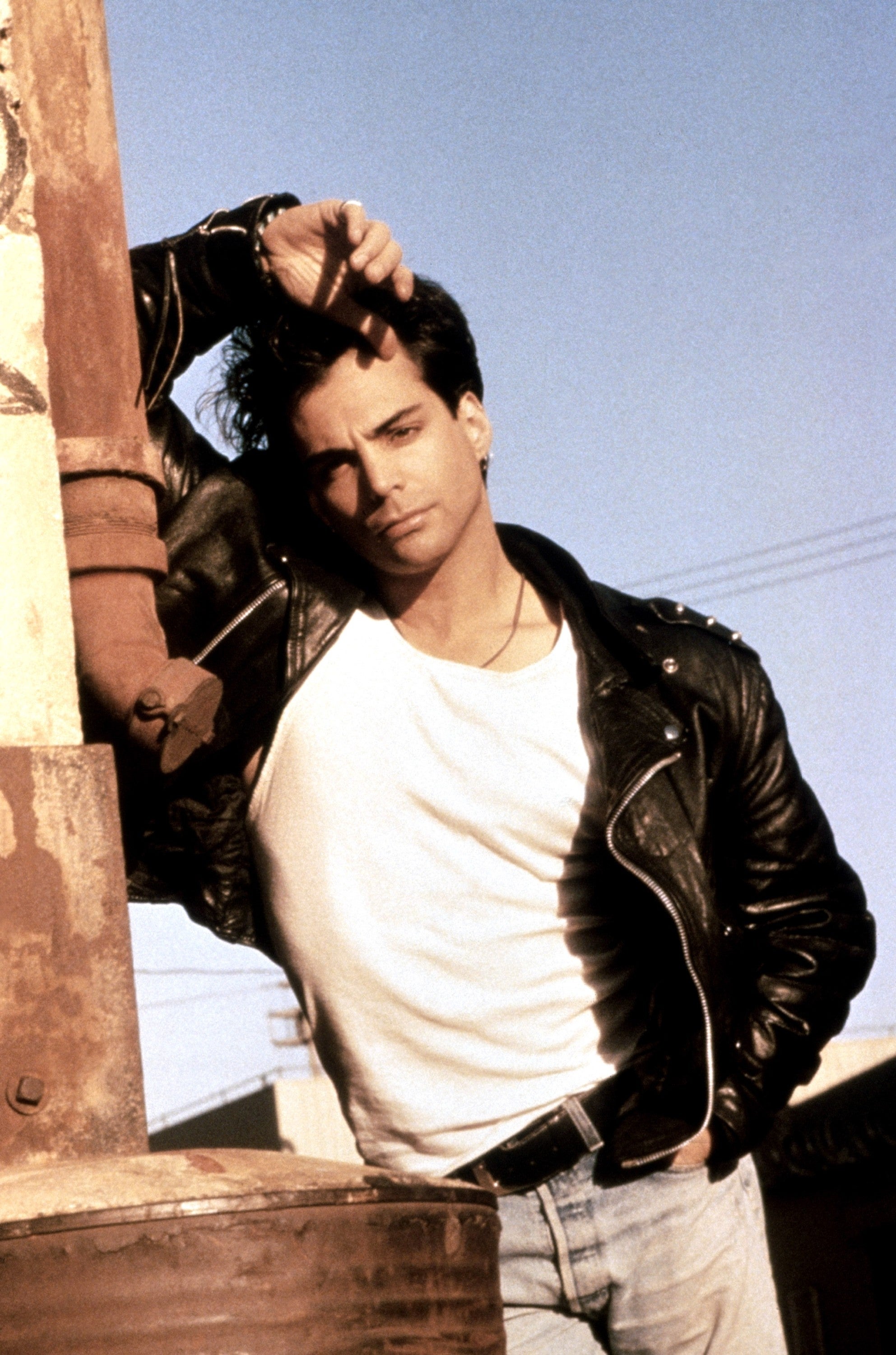 Person posing in a casual style, wearing a leather jacket and jeans, leaning against a rusted metal structure