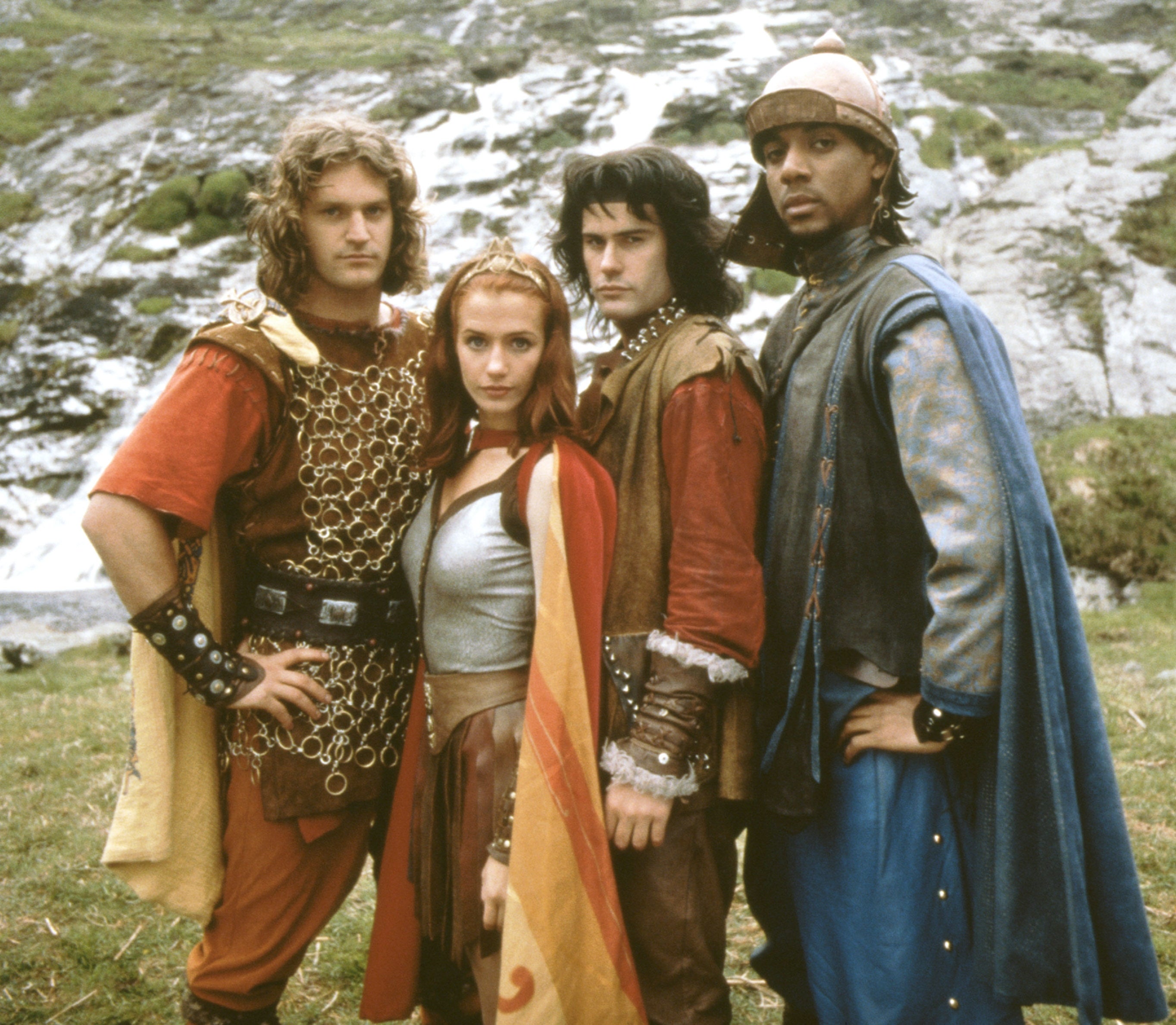 Four actors in medieval fantasy costumes stand outdoors in front of a rocky landscape, each displaying different medieval styles