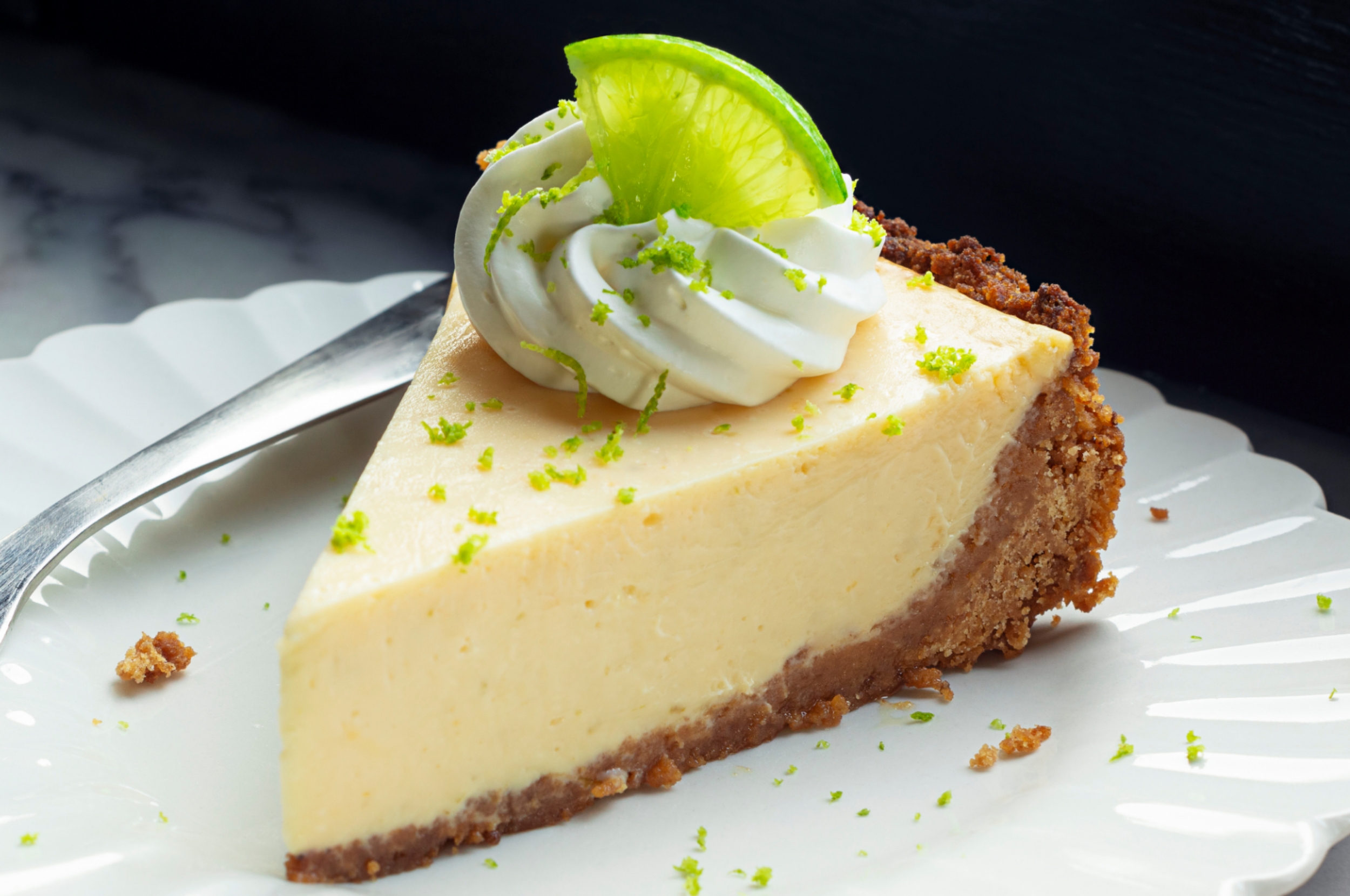 A slice of key lime pie with whipped cream and a lime garnish on a plate, with a fork beside it