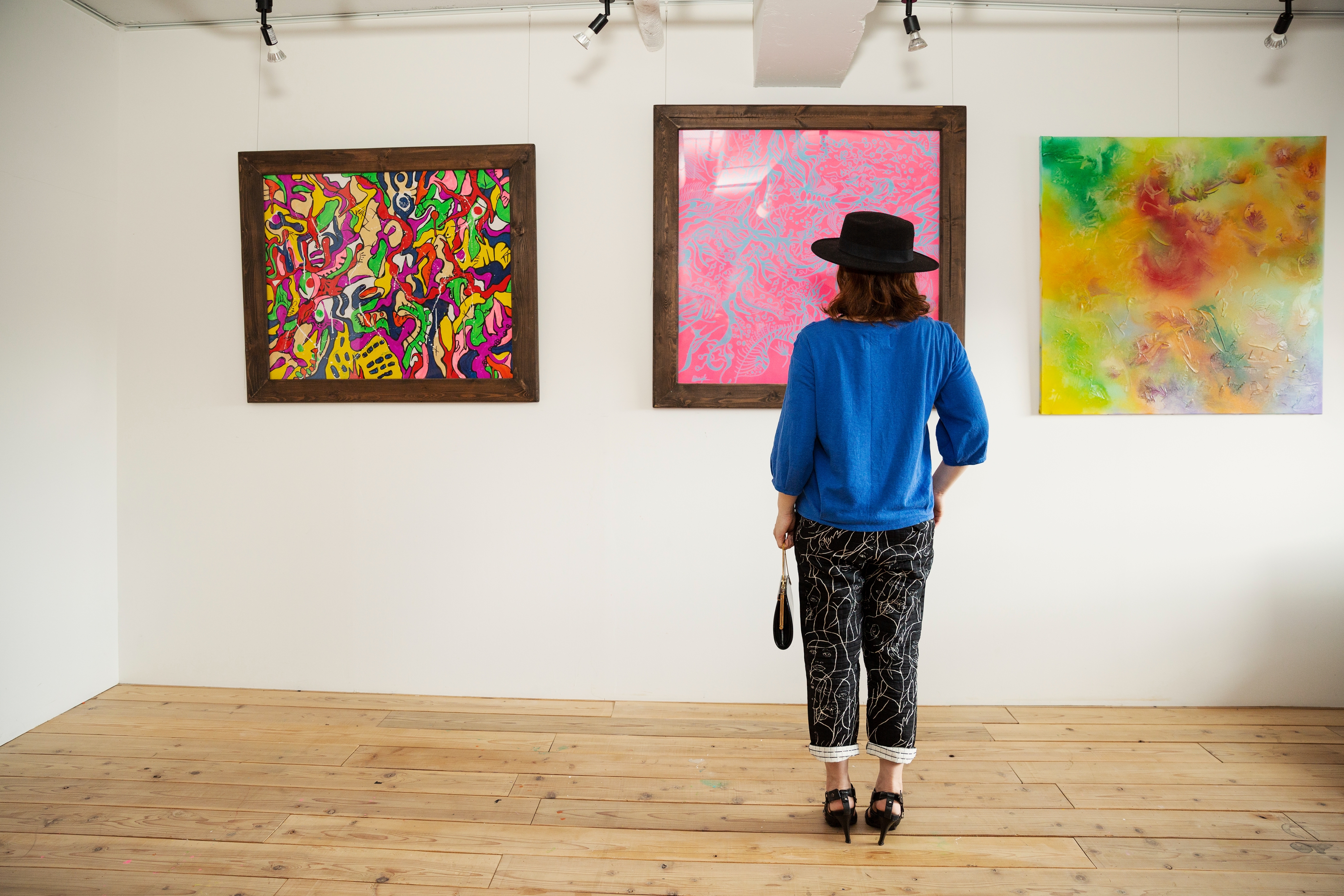 Person i mønstrede bukser og en vidt brim hatt som ser på tre abstrakte kunstverk i et galleri