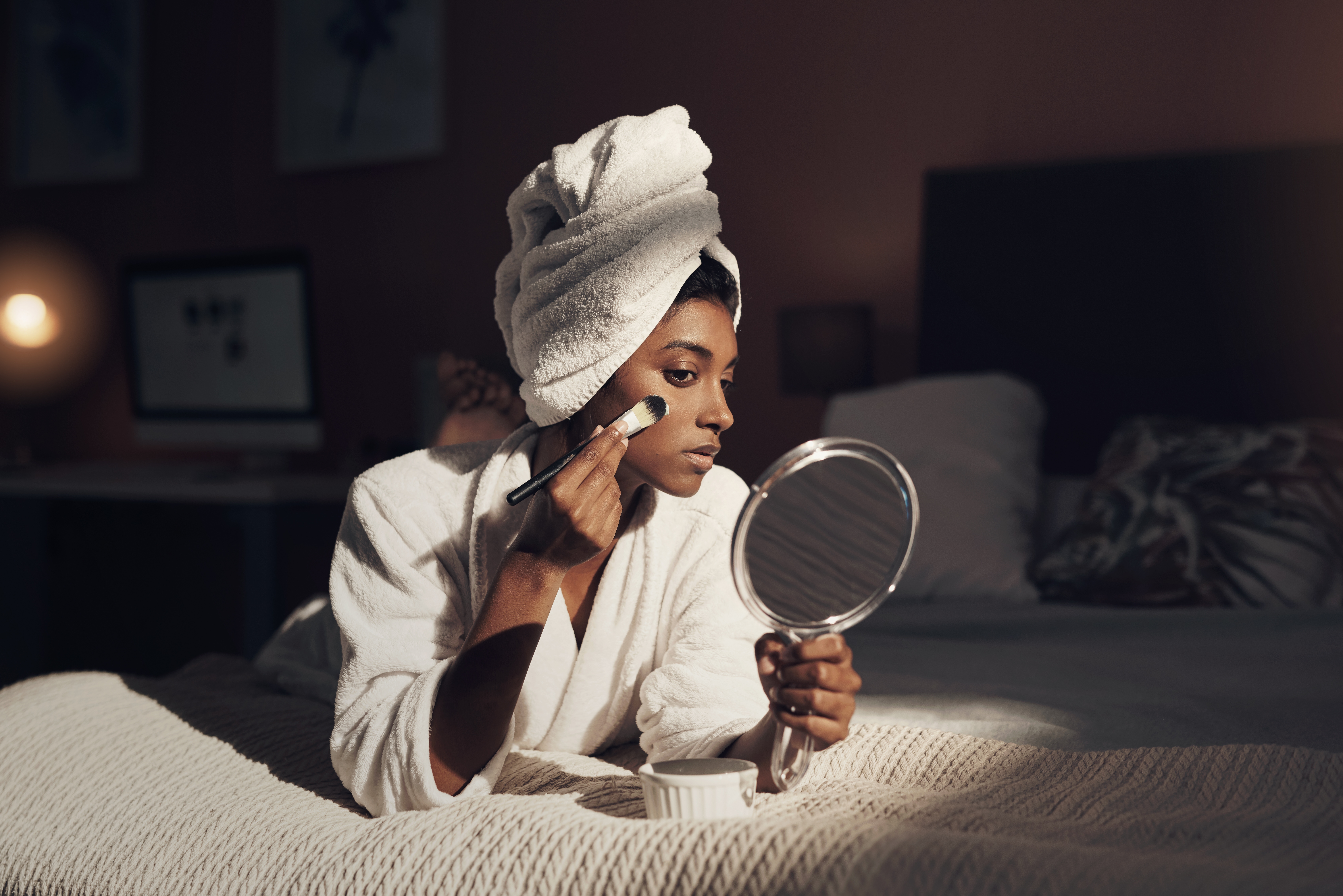 Person i en kappe, håndkle på hodet, påfør ansiktsmaske med børste mens du holder et speil, ligger på sengen i et koselig, mykt opplyst rom