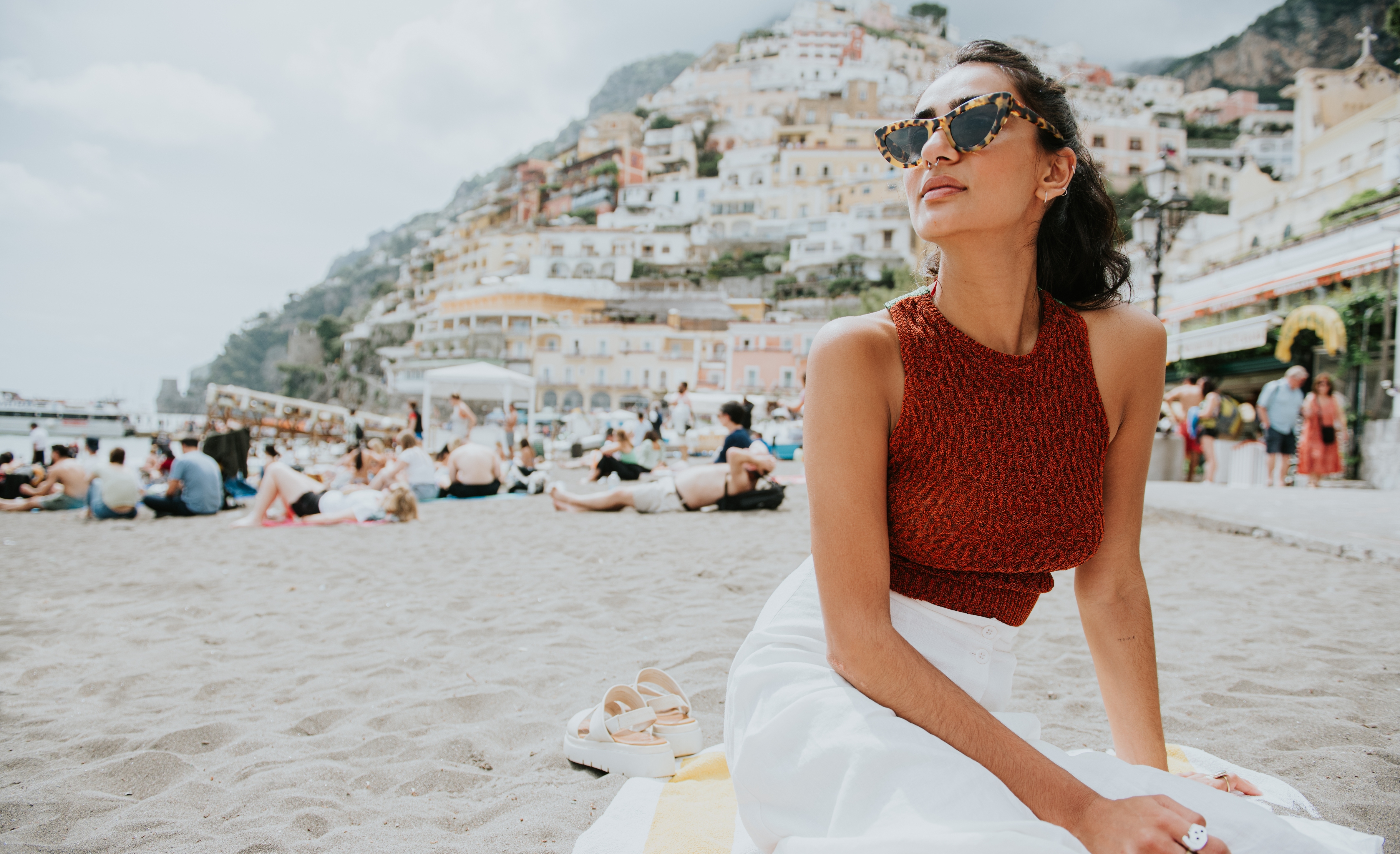 Kvinne i en ermeløs topp og langt skjørt som sitter på en sandstrand, med fargerike kystbygninger i bakgrunnen