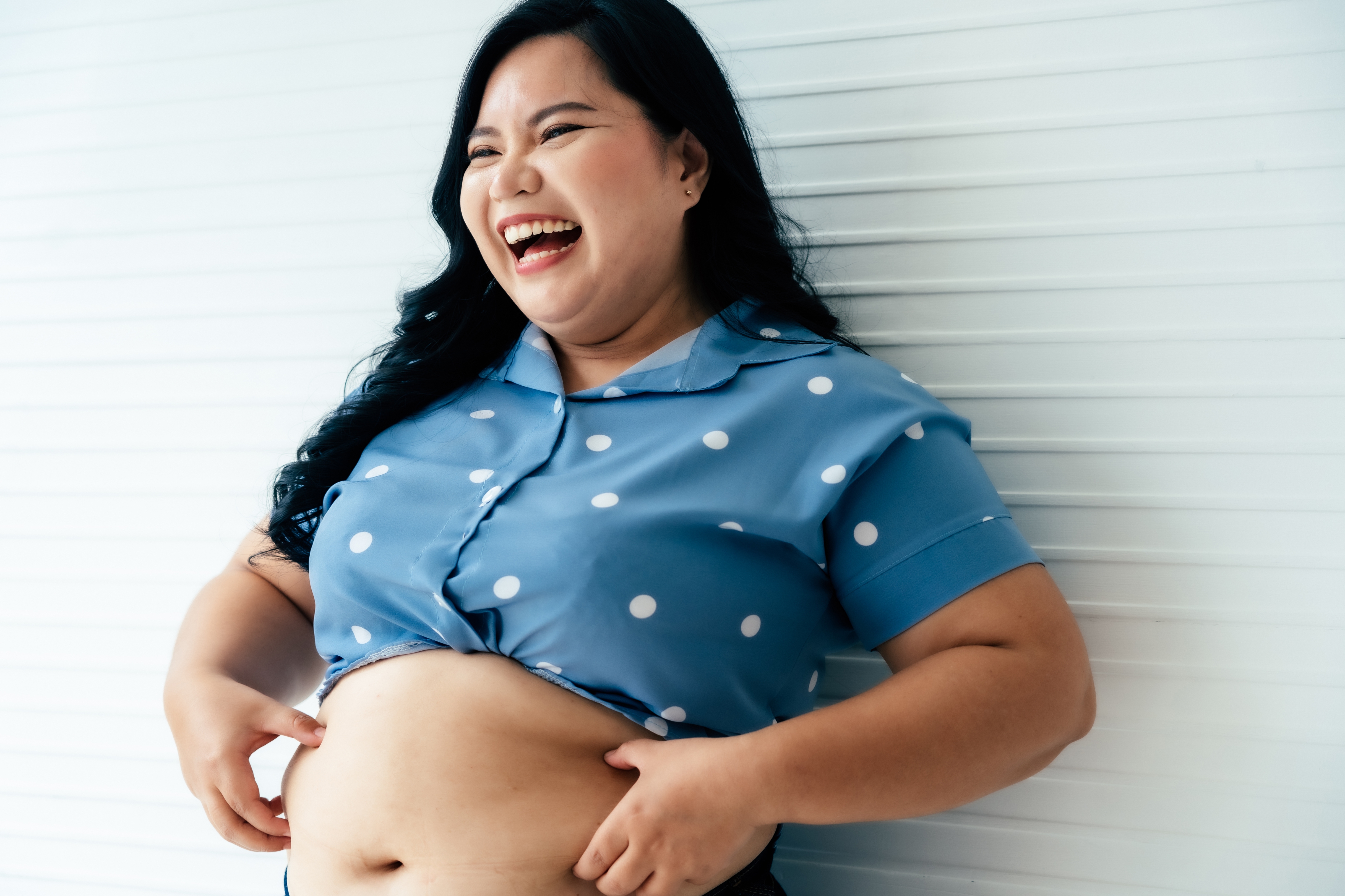 Personen poserer med glede, iført en polka-dot-topp, lekende holder midriffen. De smiler og står og står foran en hvit bakgrunn