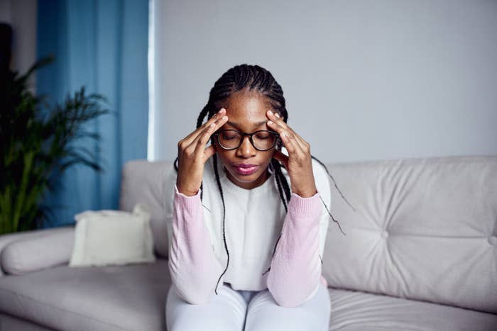 Person sitting on a couch, touching their temples, appearing deep in thought or stressed. Houseplant and pillow in the background