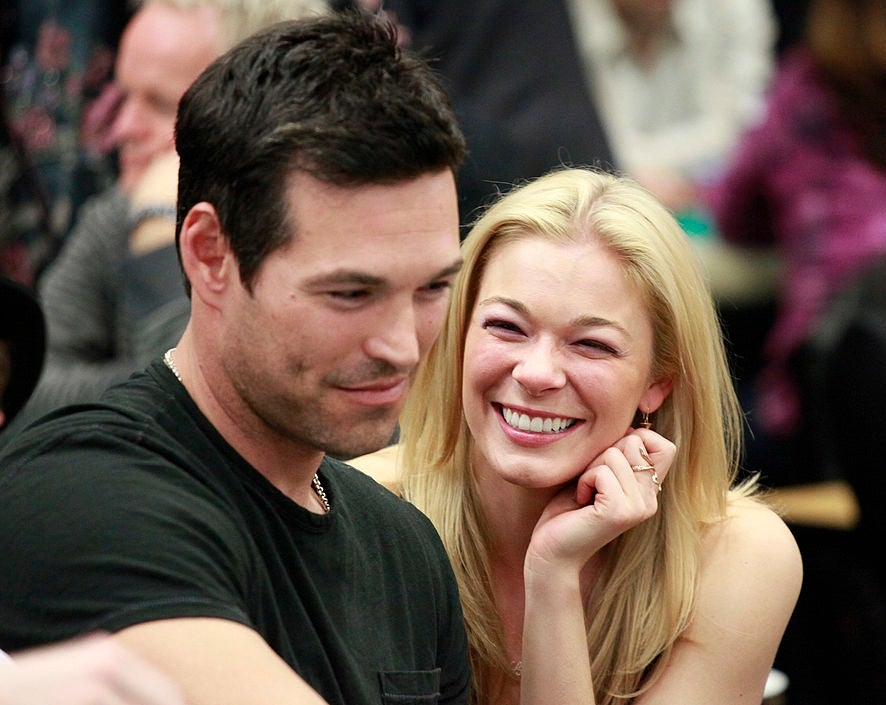A smiling woman and a man, casually dressed, sit closely at a public event. The atmosphere is relaxed and cheerful