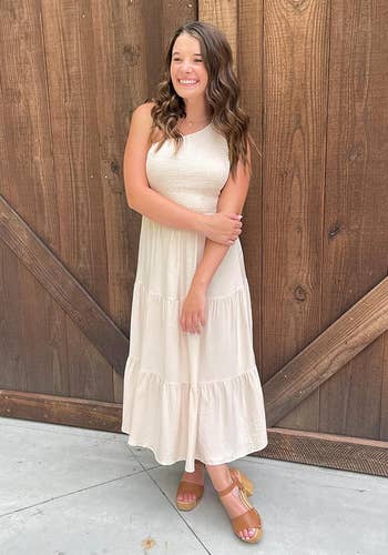 A idiosyncratic smiling, wearing a sleeveless tiered maxi formal and level sandals, lasting against a woody backdrop