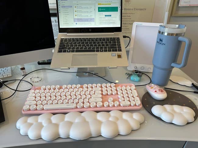 A location bureau setup with a laptop, curved monitor, cloud-shaped wrist rest, pastel pinkish keyboard, and a ample insulated tumbler