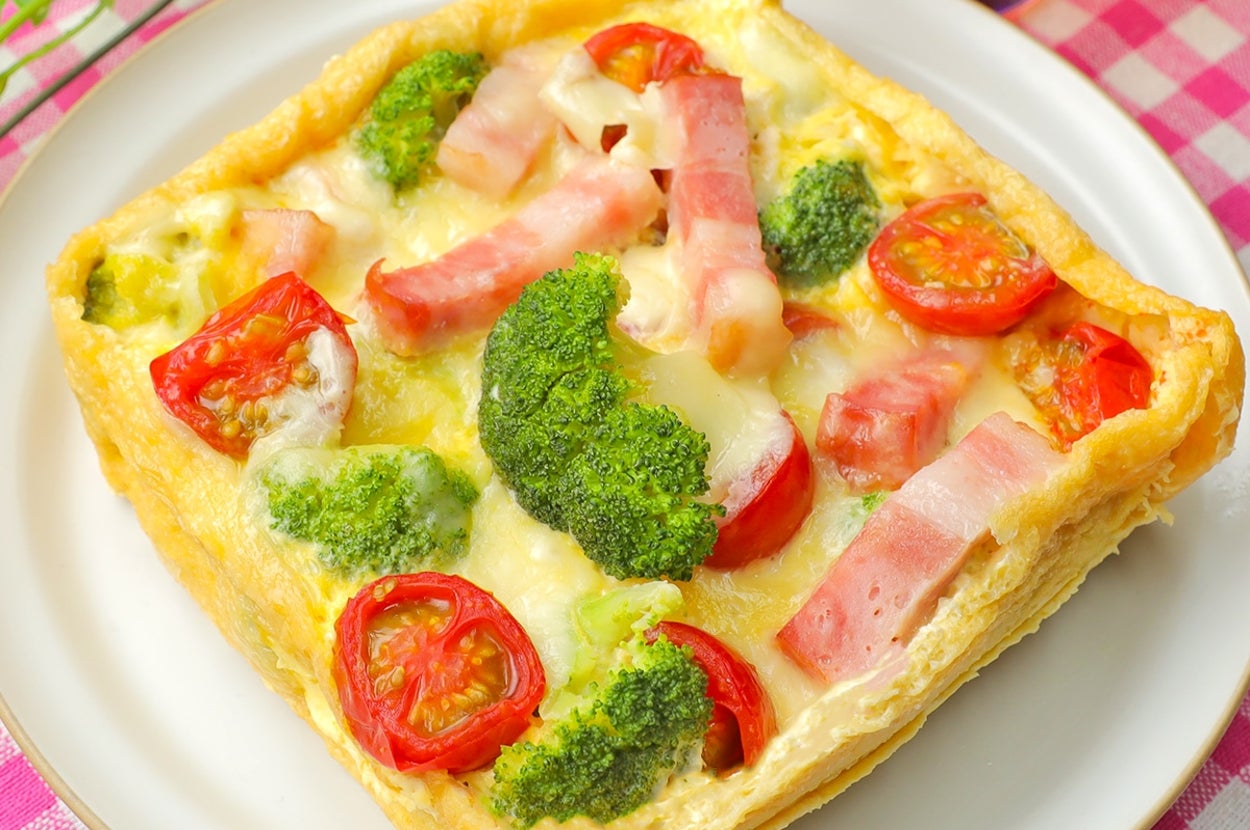 ブロッコリー、トマト、ベーコンがのった四角いキッシュが白い皿に載っている。後ろに飲み物の入ったグラス。