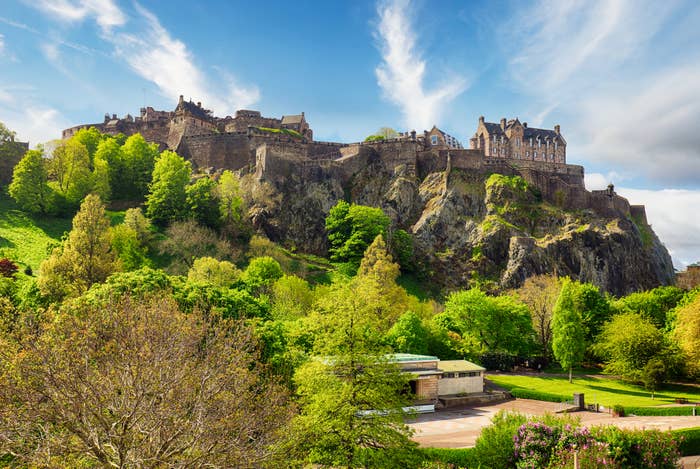 View of Edinburgh Castle atop a rocky hill, surrounded by lush greenery and trees under a partly cloudy sky