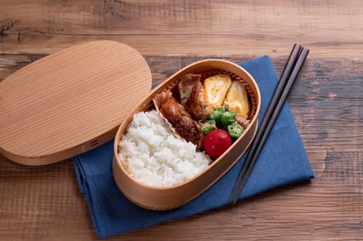 弁当箱の中に、ご飯、卵焼き、鶏肉、オクラ、ミニトマトが入っており、青い布の上に置かれている。