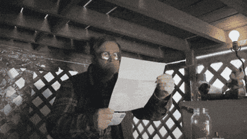 Man under a wooden structure squints at a paper, adjusting his glasses and focusing intently