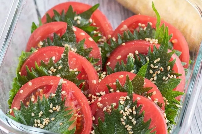 透明容器にトマトとシソの葉が盛り付けられ、ゴマが散らされています。背景にパンがあります。
