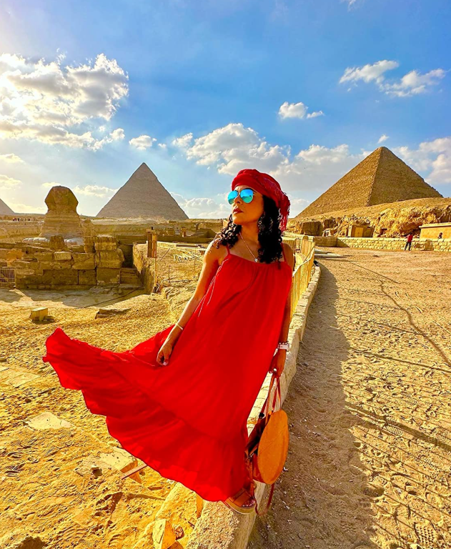 Person successful  a flowing reddish  formal  and reddish  chapeau  poses stylishly by the pyramids, holding a circular  bag