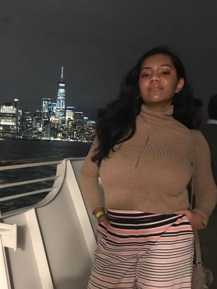 Person posing on a boat, wearing a turtleneck sweater and striped pants, with a city skyline illuminated in the background at night