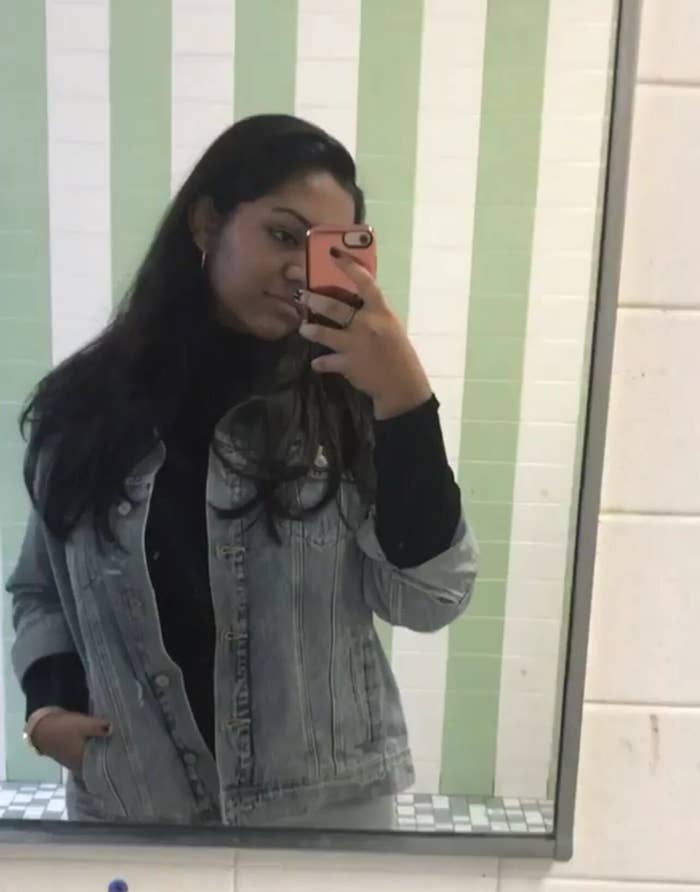 Person taking a mirror selfie in a restroom, wearing a denim jacket over a black top, holding a pink phone