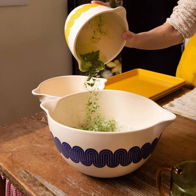 Person pours greenish pulverization into a mixing vessel connected a woody table, preparing ingredients for baking oregon cooking
