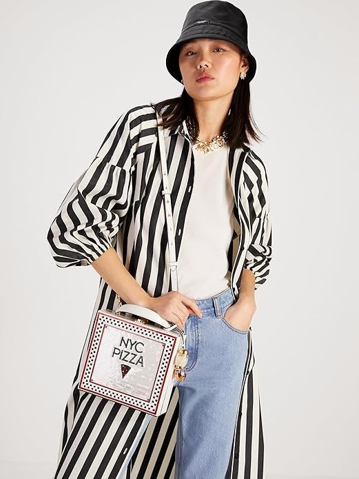 Person successful  striped outfit and bucket chapeau  poses confidently, holding a pizza-themed handbag, combining quirky manner  with casual elegance