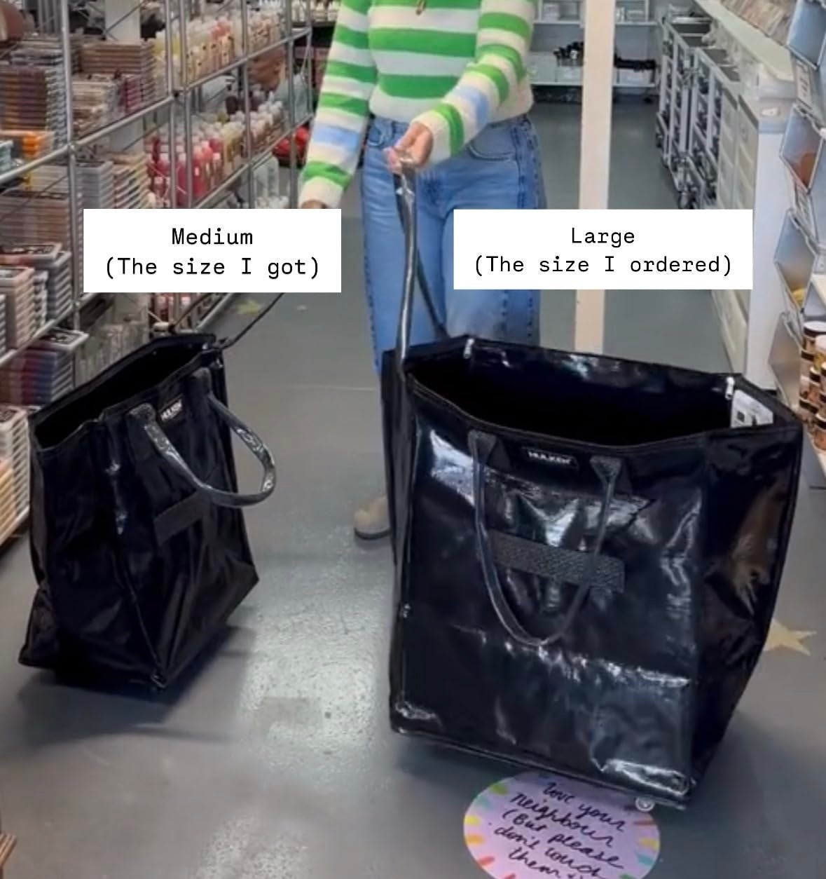 Person in striped sweater comparing two black tote bags: medium (size received) and large (size ordered) in a store aisle