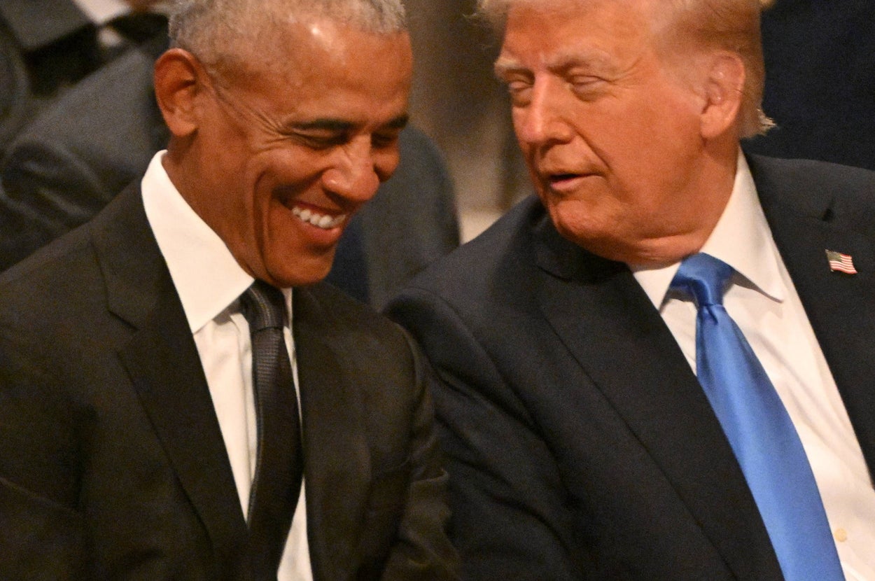 Two men in suits, one in a black suit, the other in navy with a blue tie, are seated and appear to be sharing a light-hearted conversation