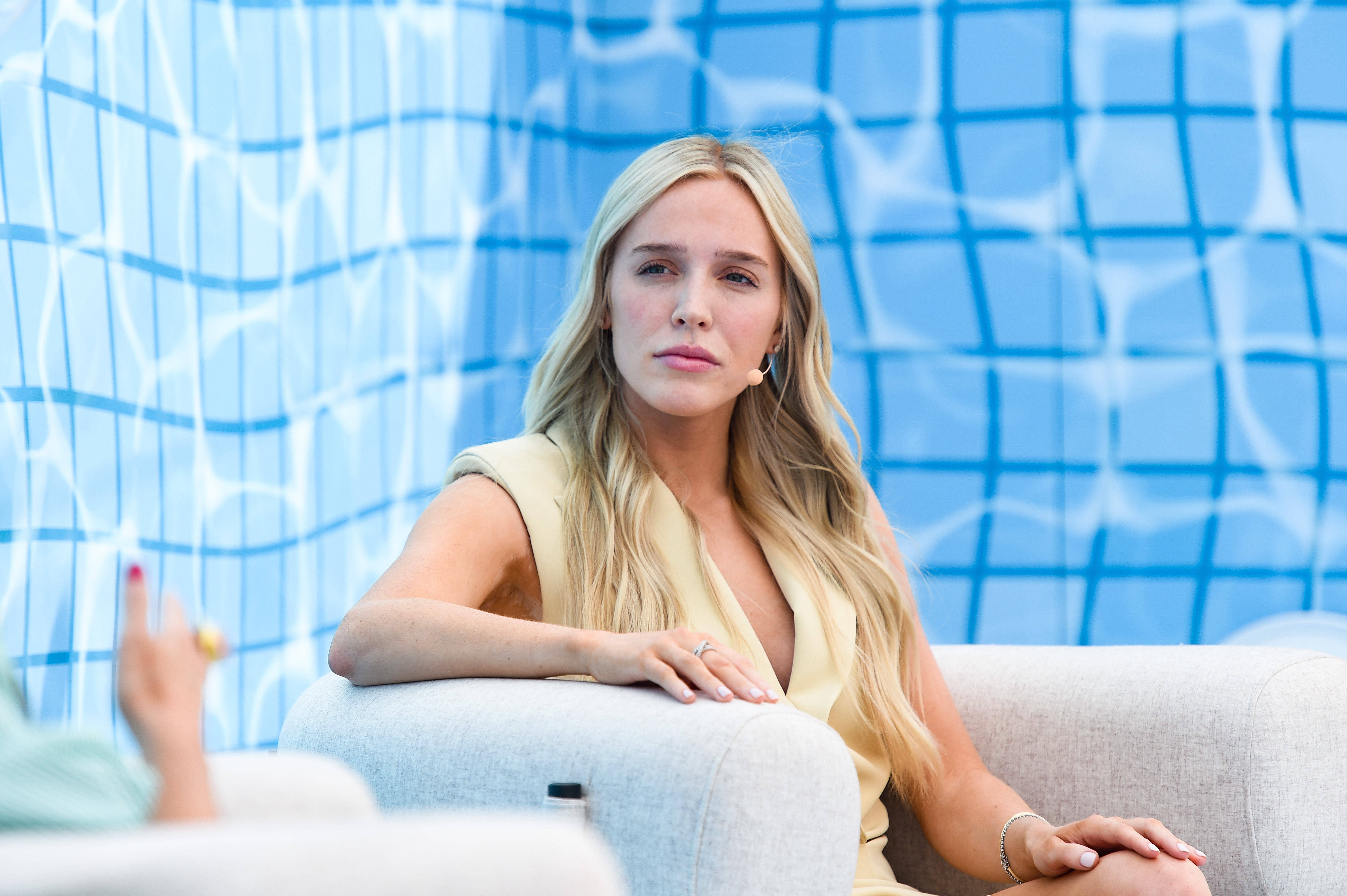 Alex Cooper seated on a light sofa, wearing a stylish sleeveless top, against a geometric patterned backdrop